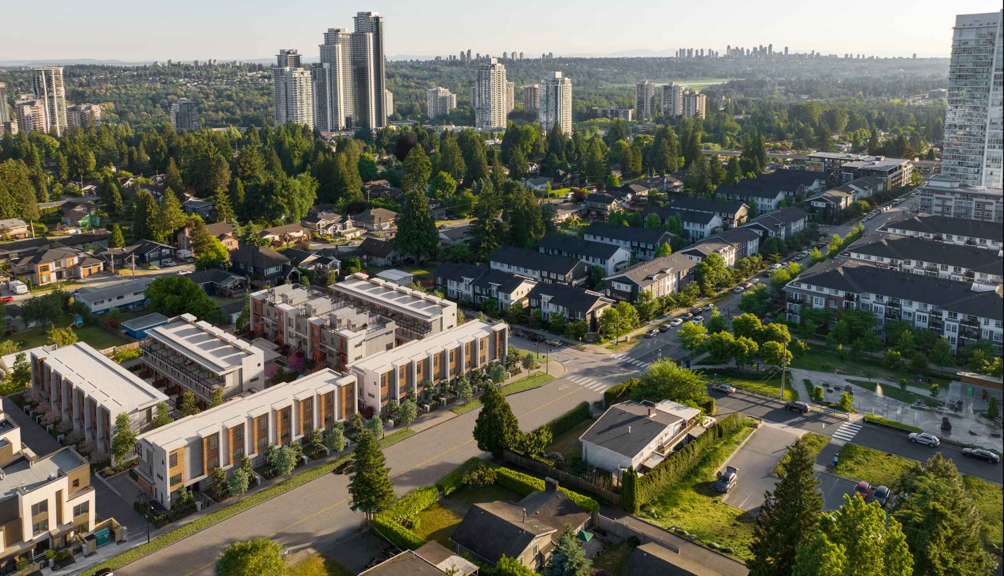 The Grove – An aerial drone view of a new townhome complex under a white rooftop, situated among a suburban neighborhood of houses and low-rise buildings with high-rise towers and a city skyline in the distance.