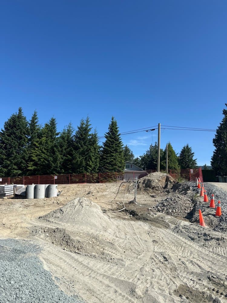 Tunbridge Crossing – Early construction site photo with dirt mounds, concrete drainage rings, orange safety fencing, and traffic cones, evergreen trees behind.