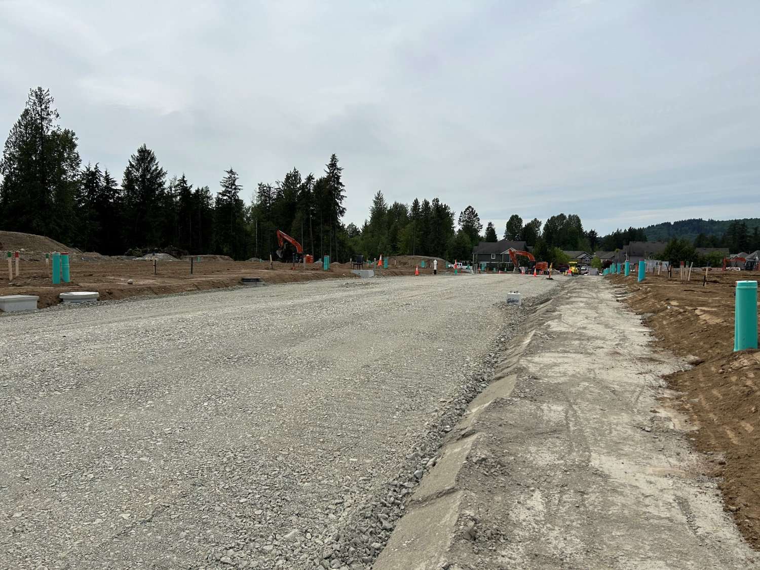 Tunbridge Crossing – New gravel road under construction with an excavator and a freshly poured concrete curb/sidewalk section, homes visible in the distance.