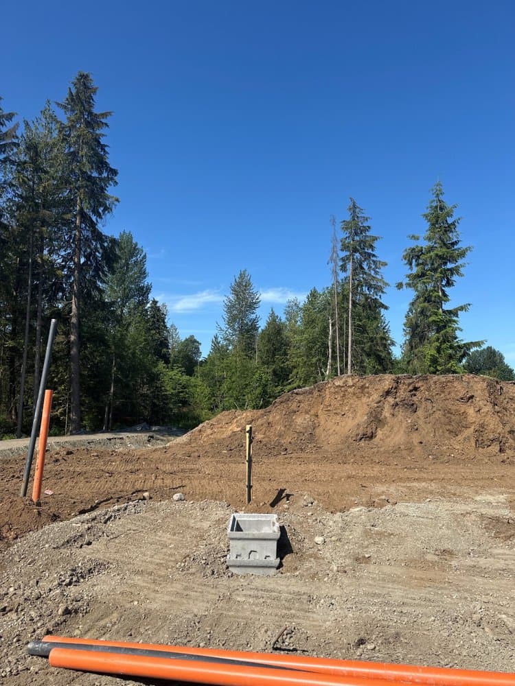 Tunbridge Crossing – Bare construction lot with a concrete utility box, orange conduit pipes, and a dirt mound, forest in the background.