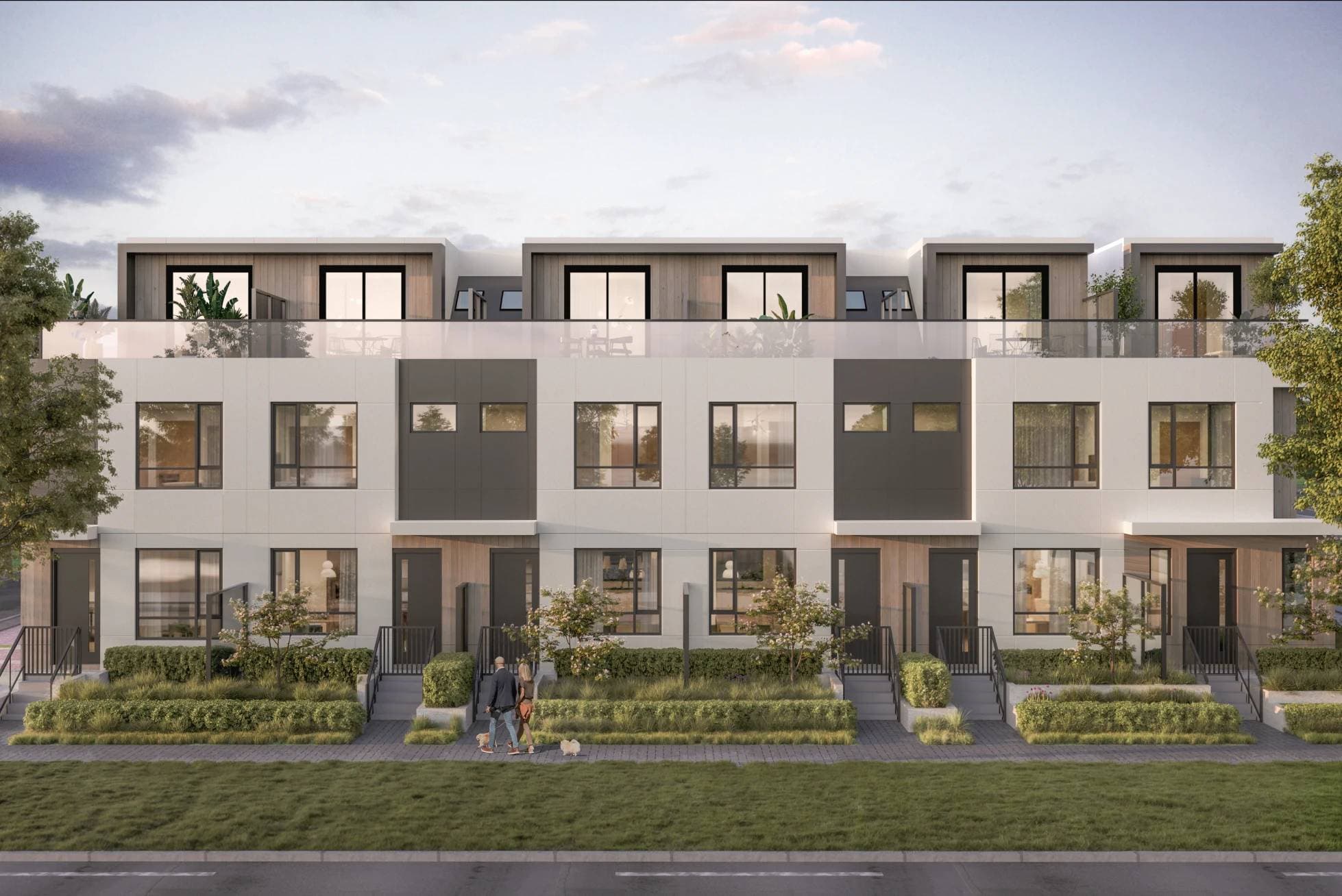 Emera – A row of modern three-story townhomes features white and charcoal cladding with wood-look accents and glass-railed rooftop patios under a soft dusk sky. Landscaped walkways, small garden beds, and short steps lead to private front entrances where two people walk a dog along the sidewalk.