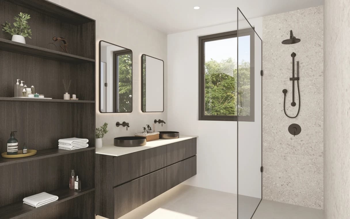 Emera – A modern bathroom featuring a dark wood double vanity with matte black vessel sinks, backlit rounded mirrors, and built-in open shelving on the left. On the right, a walk-in shower is enclosed by a glass panel and finished with terrazzo-style wall tiles and matte black fixtures, with daylight streaming in through a window overlooking lush green trees.