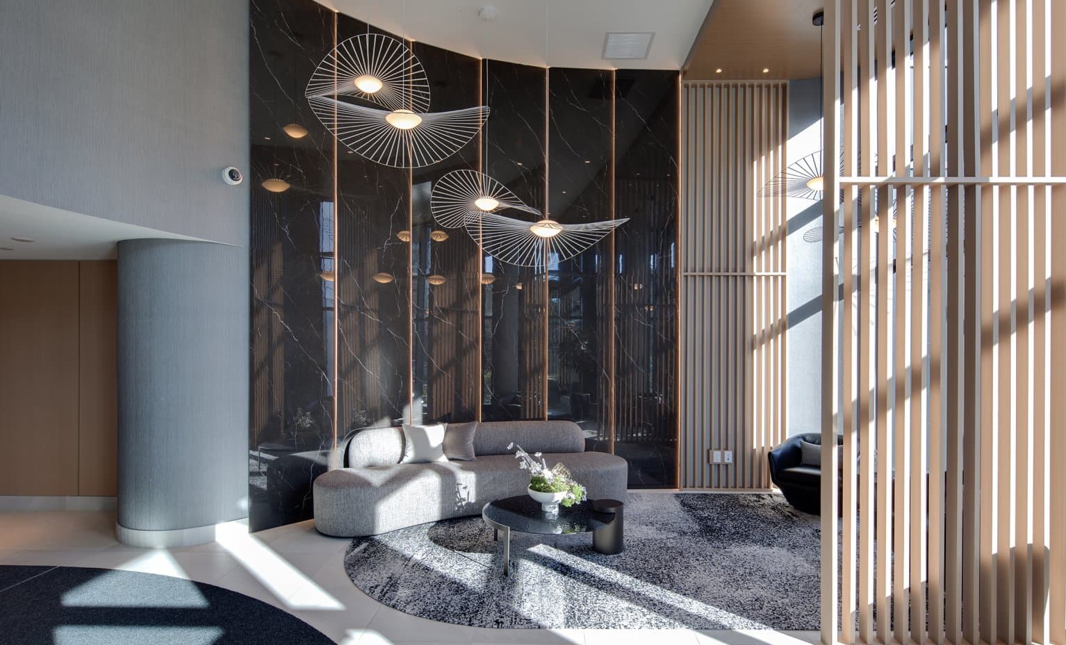 Wordsworth – A modern, sunlit lobby lounge featuring a curved gray sofa, round coffee table, and an accent rug against a polished black marble feature wall. Distinctive wire-frame pendant lights hang from above, complemented by vertical light-wood slat partition screens and bright sunlight casting geometric shadows.