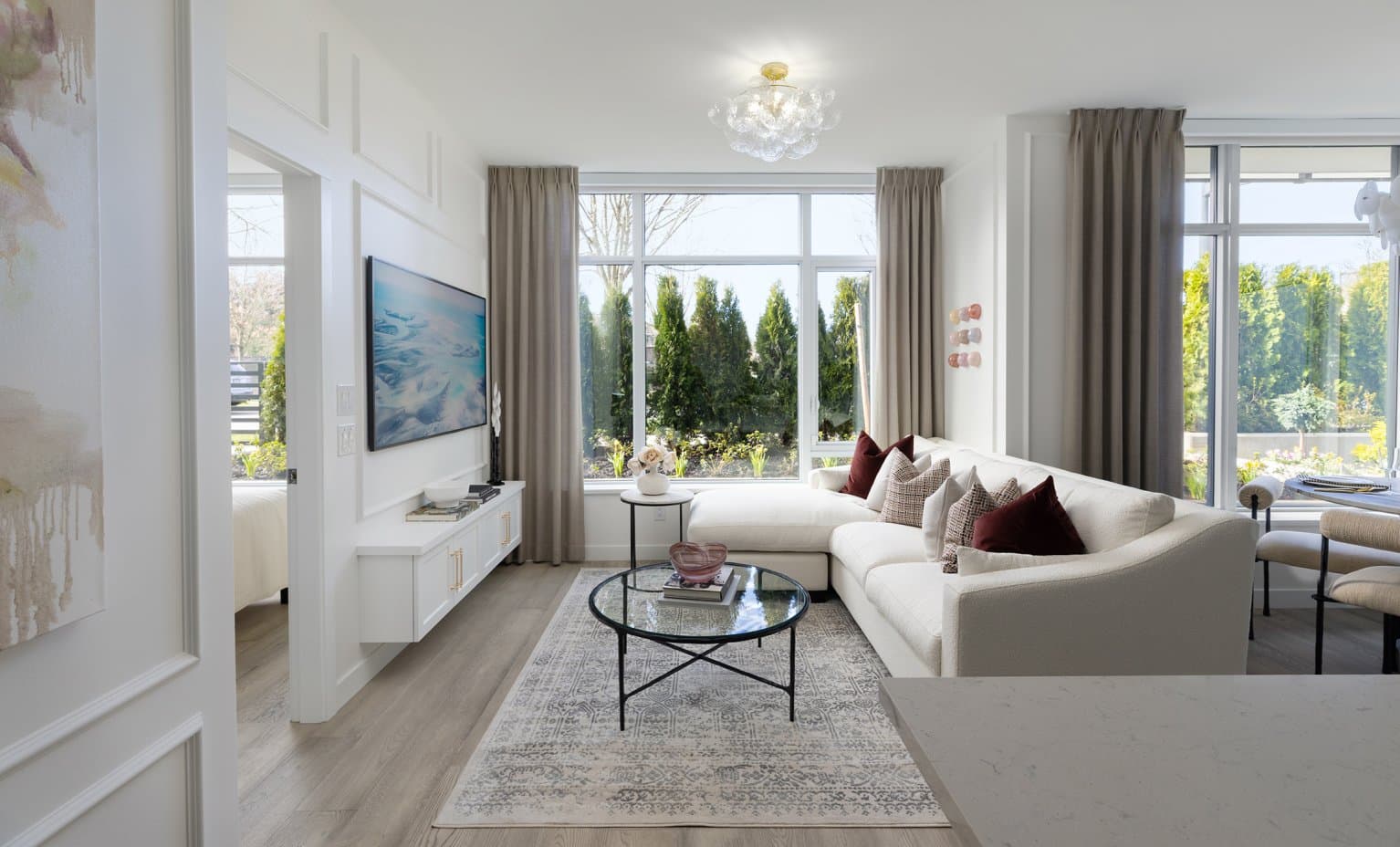 Wordsworth – A bright, elegant living room featuring a cream-colored sectional sofa, round glass coffee table, and a wall-mounted TV over a floating white media console. Large floor-to-ceiling windows with taupe drapery look out onto lush evergreen trees, filling the space with natural daylight.