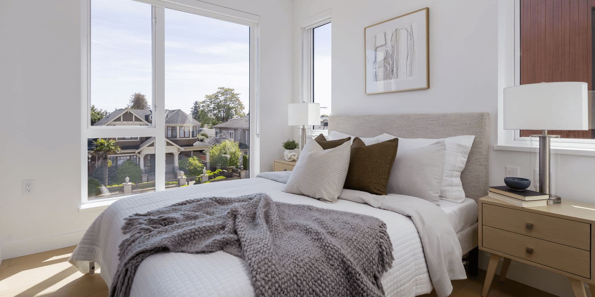BE 49th – Bedroom with a grey upholstered headboard, layered white and knit throw bedding, and floor-to-ceiling windows looking out over neighboring rooftops and trees.