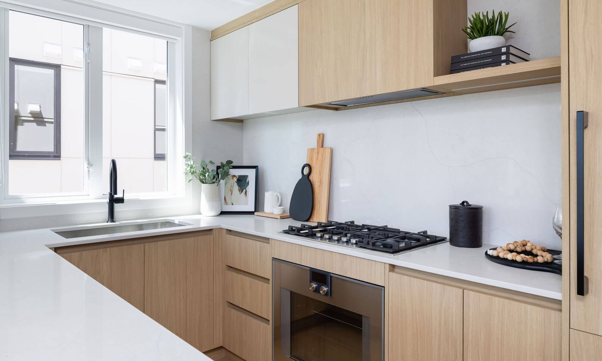 BE 49th – Close-up of a light wood kitchen counter with a gas cooktop, black kettle, cutting boards, and a marble backsplash beside a window.