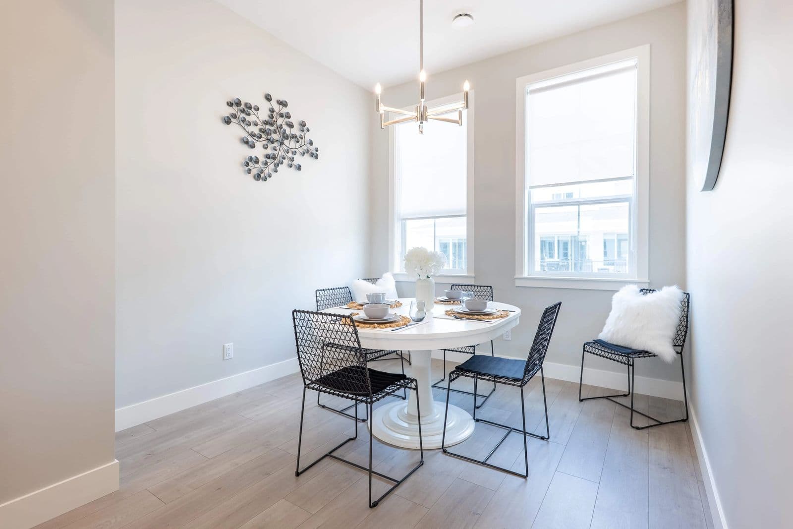 Cedarbrook – A dining area with a round white table, black wire chairs, a decorative metal wall sculpture, and a pendant light fixture.