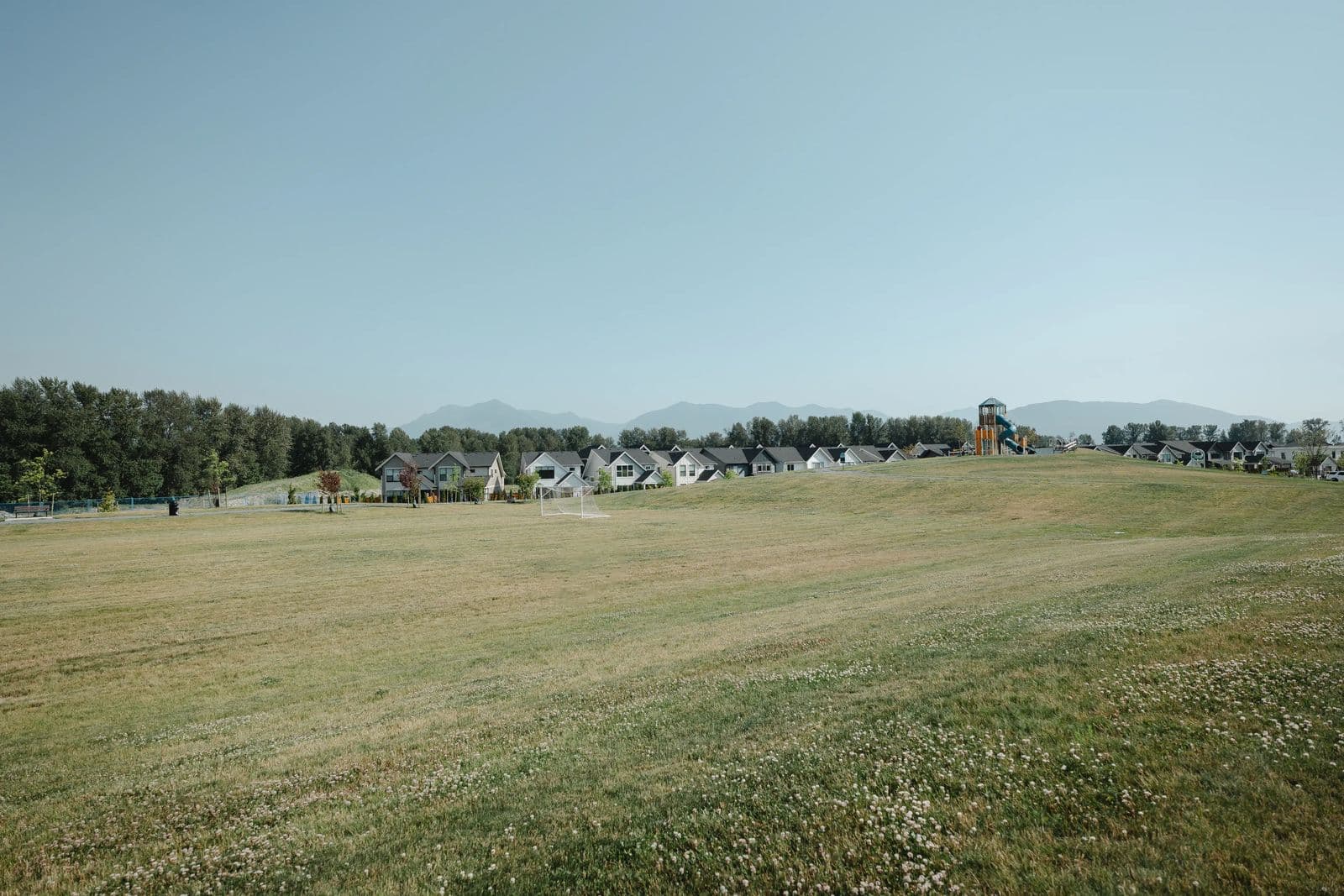 Cedarbrook – An open grass field with a soccer net and a playground tower, with a row of townhomes and mountains in the distance.
