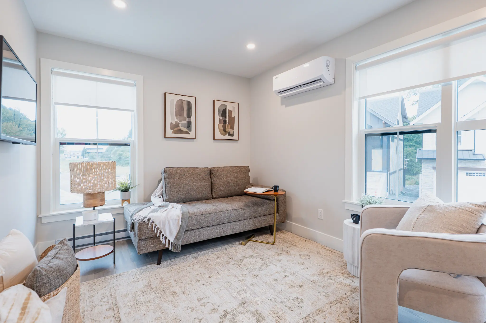 Cedarbrook – A cozy den with a grey loveseat, a wall-mounted air conditioning unit, framed abstract art, and a second seating area with a beige armchair.
