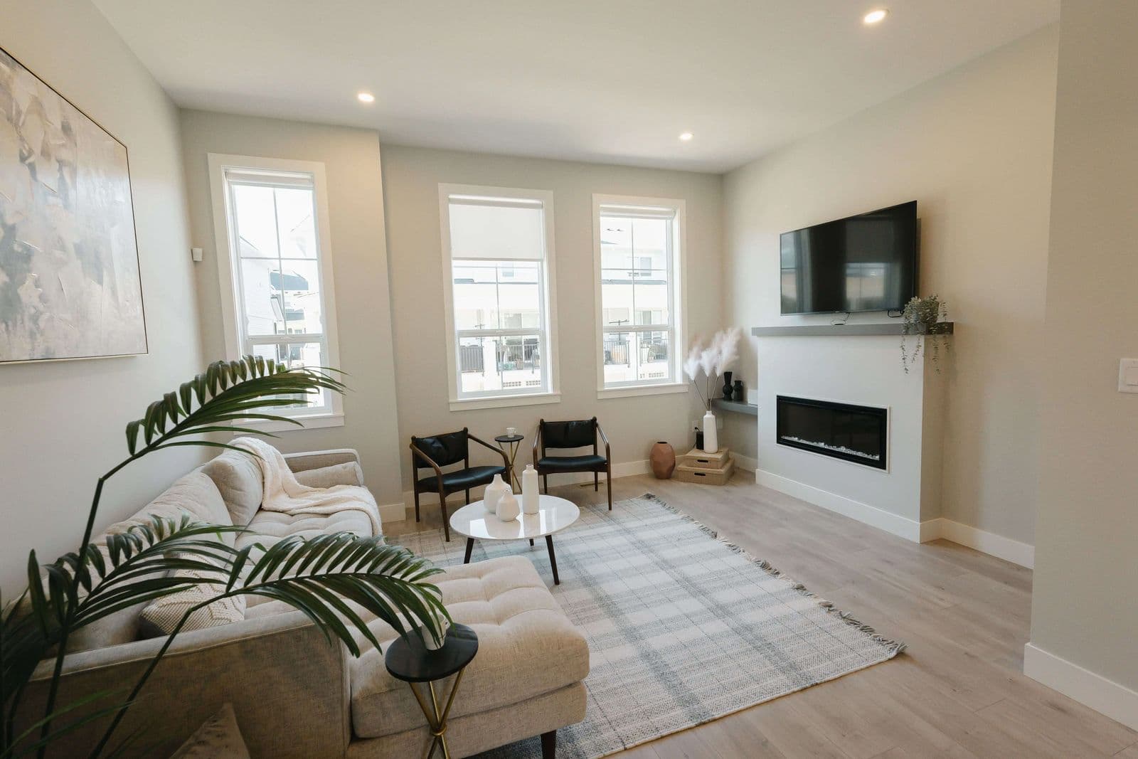 Cedarbrook – A living room with a light grey sofa and ottoman, an electric fireplace under a mounted TV, and three large windows.