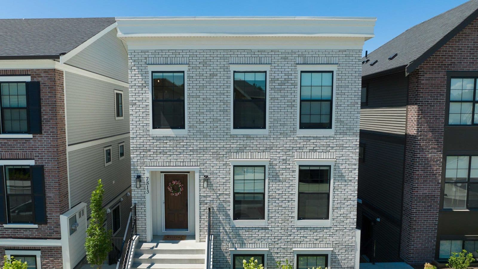 Cedarbrook – Front view of a narrow three-story home with light grey brick siding, a wood front door with a wreath, and street number 7813.