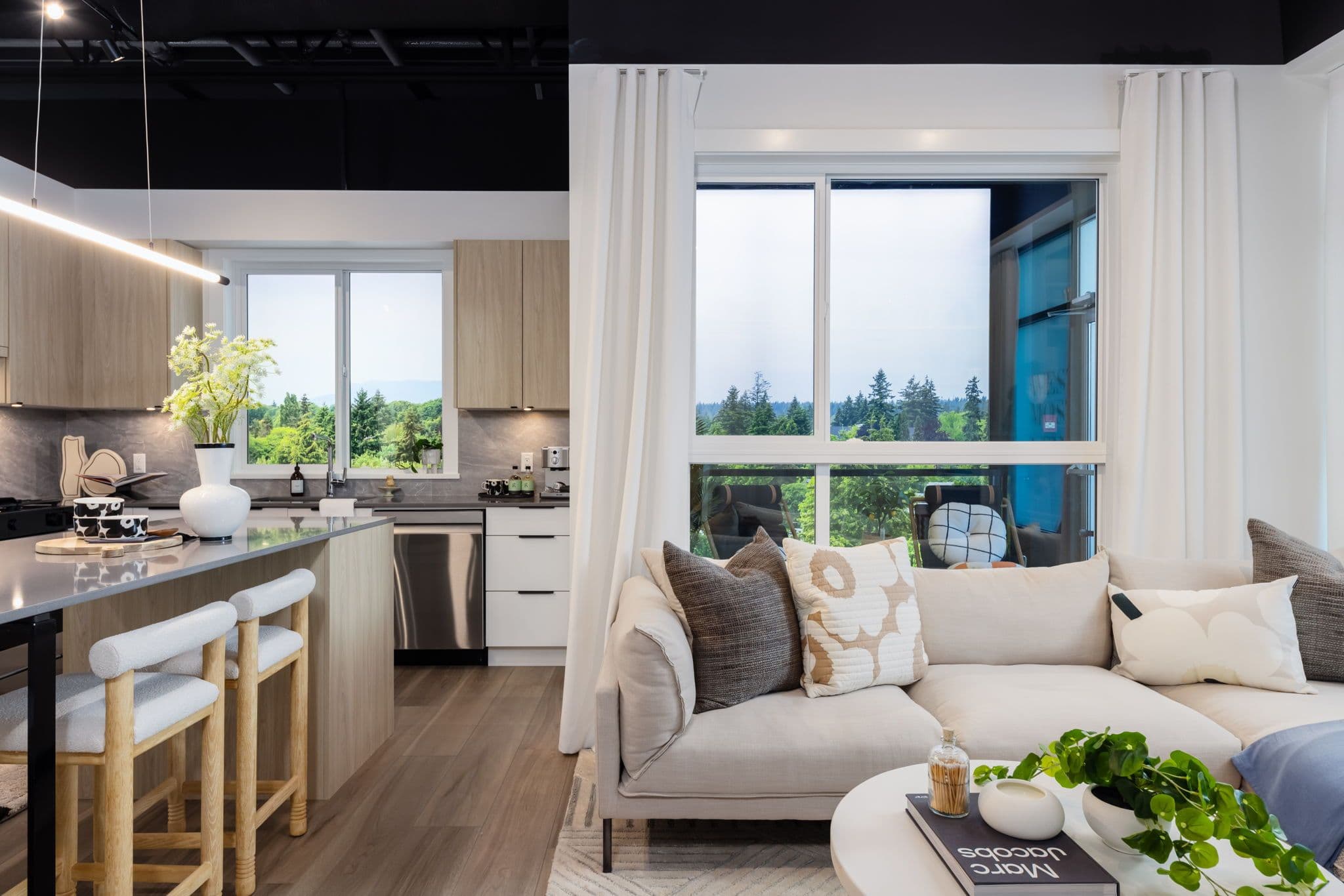 Highstreet Village – Open-concept show-suite kitchen and living room with a wood island, bar stools, beige sectional sofa, and large windows showing trees outside.