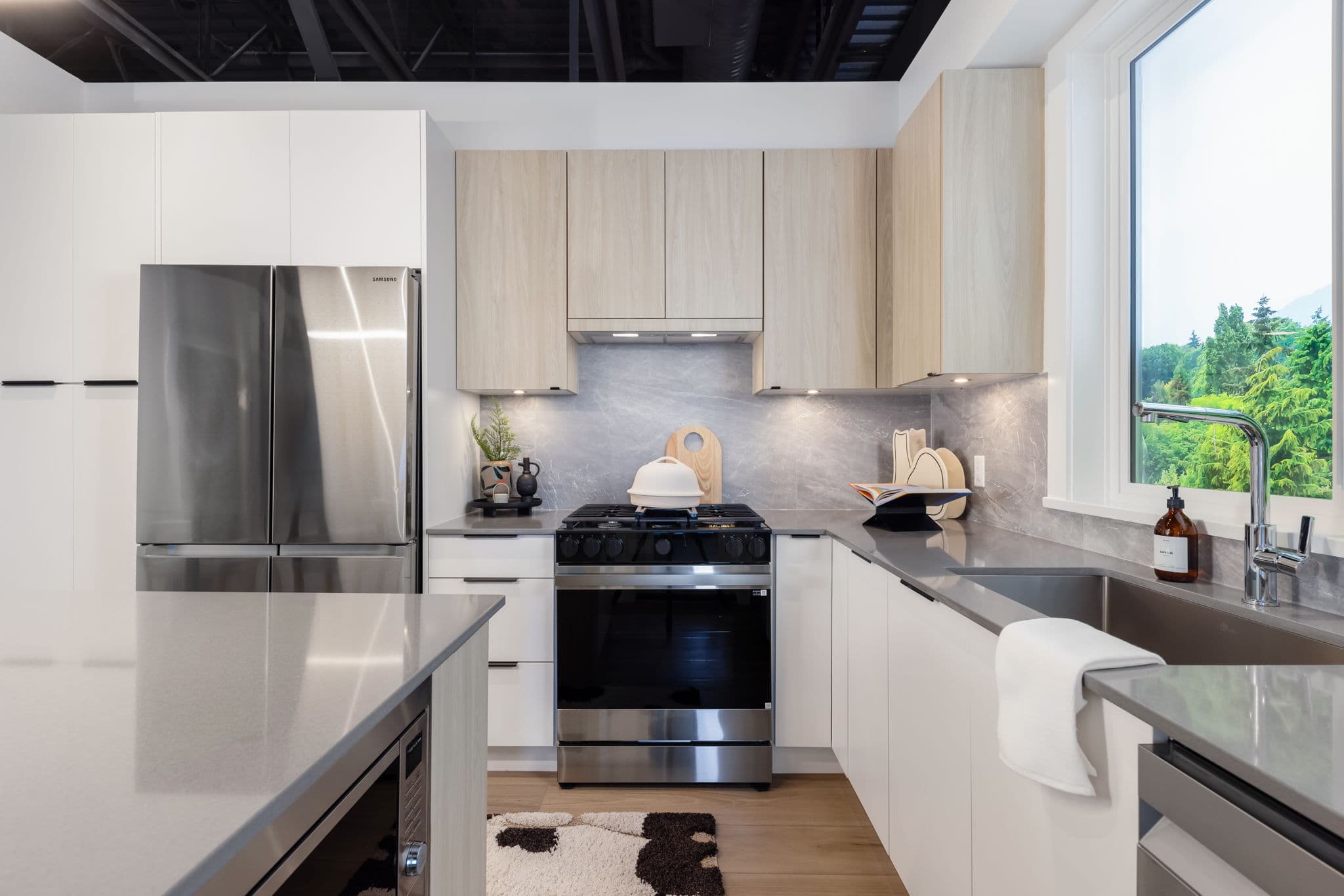 Highstreet Village – Show-suite kitchen with white and light wood cabinetry, stainless steel refrigerator and gas range, gray countertops, and a window looking out to trees.