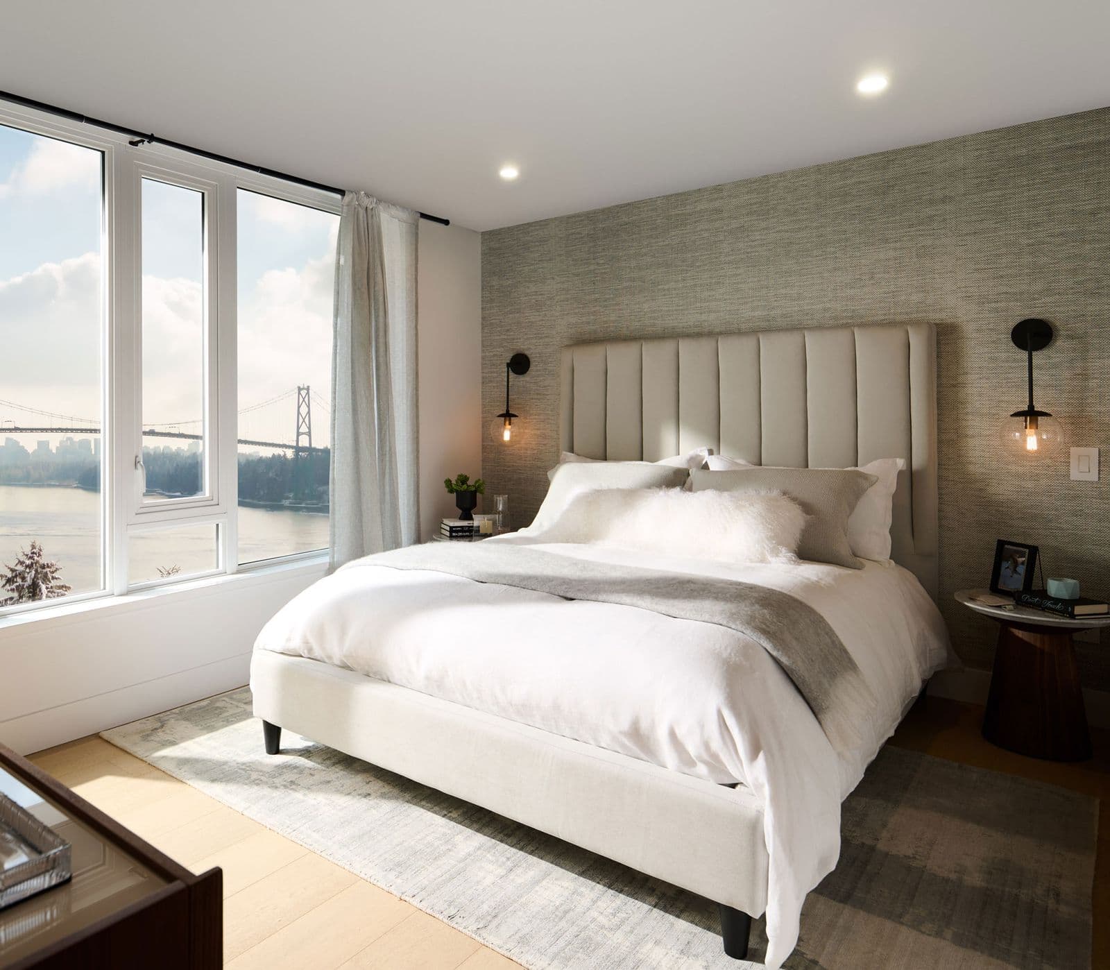 Executive on the Park – Bedroom with an upholstered bed, textured accent wall, and large windows showing a bridge and water view.