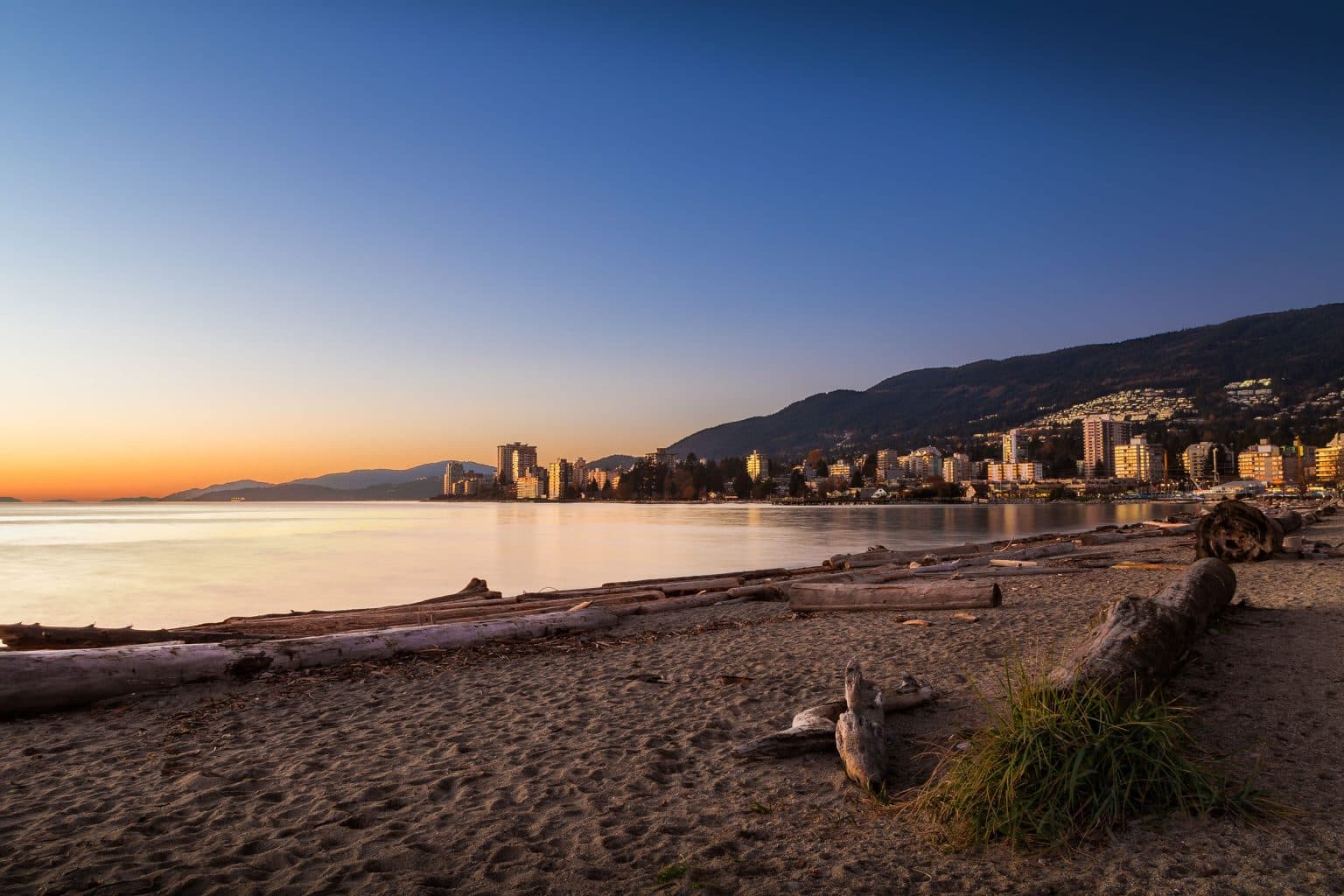 Executive on the Park – Sandy beach at sunset with driftwood logs in the foreground and a city skyline and mountains along the shoreline in the distance.