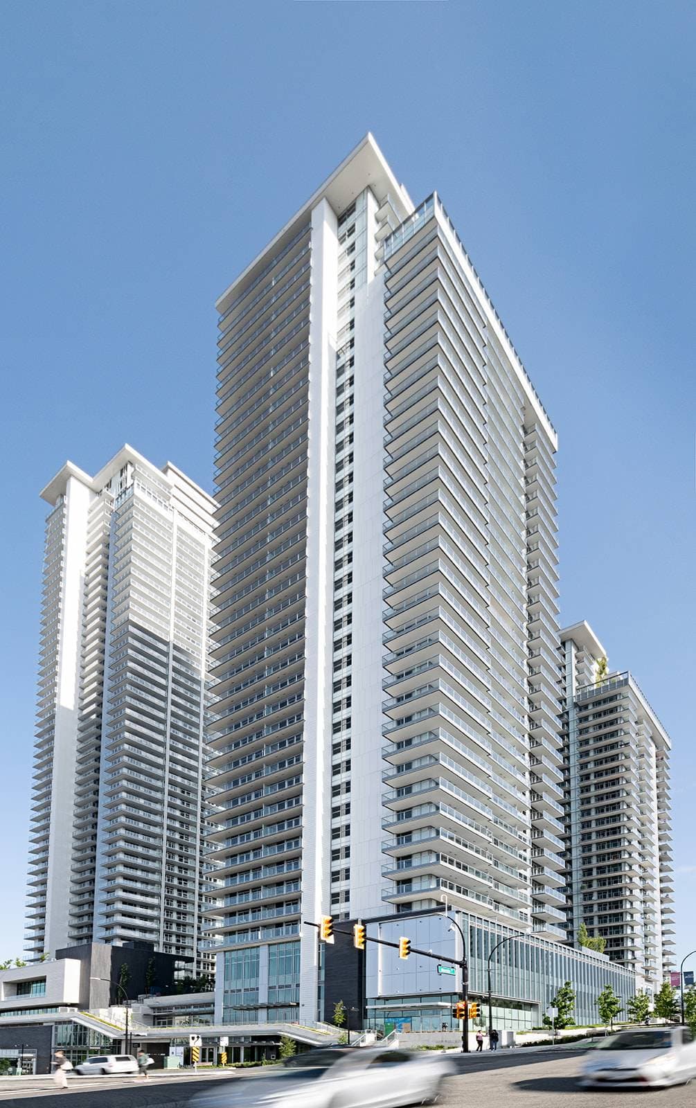 The City of Lougheed – A ground-level, upward-angled view of a modern high-rise residential tower featuring a sleek white facade, wrap-around glass balconies, and an angular roofline set against a clear blue sky. Additional matching high-rise towers and a multi-level commercial podium border a city street with traffic below.