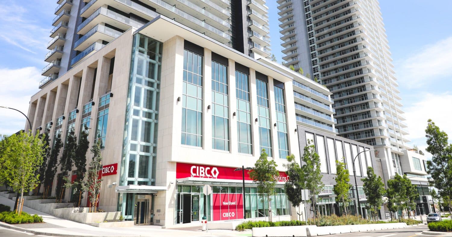 The City of Lougheed – A ground-level street view of a modern multi-story podium and high-rise residential towers, featuring a streetfront commercial space occupied by a CIBC bank branch with large glass windows and light stone cladding. Young trees and sidewalks frame the front of the complex under clear daylight.