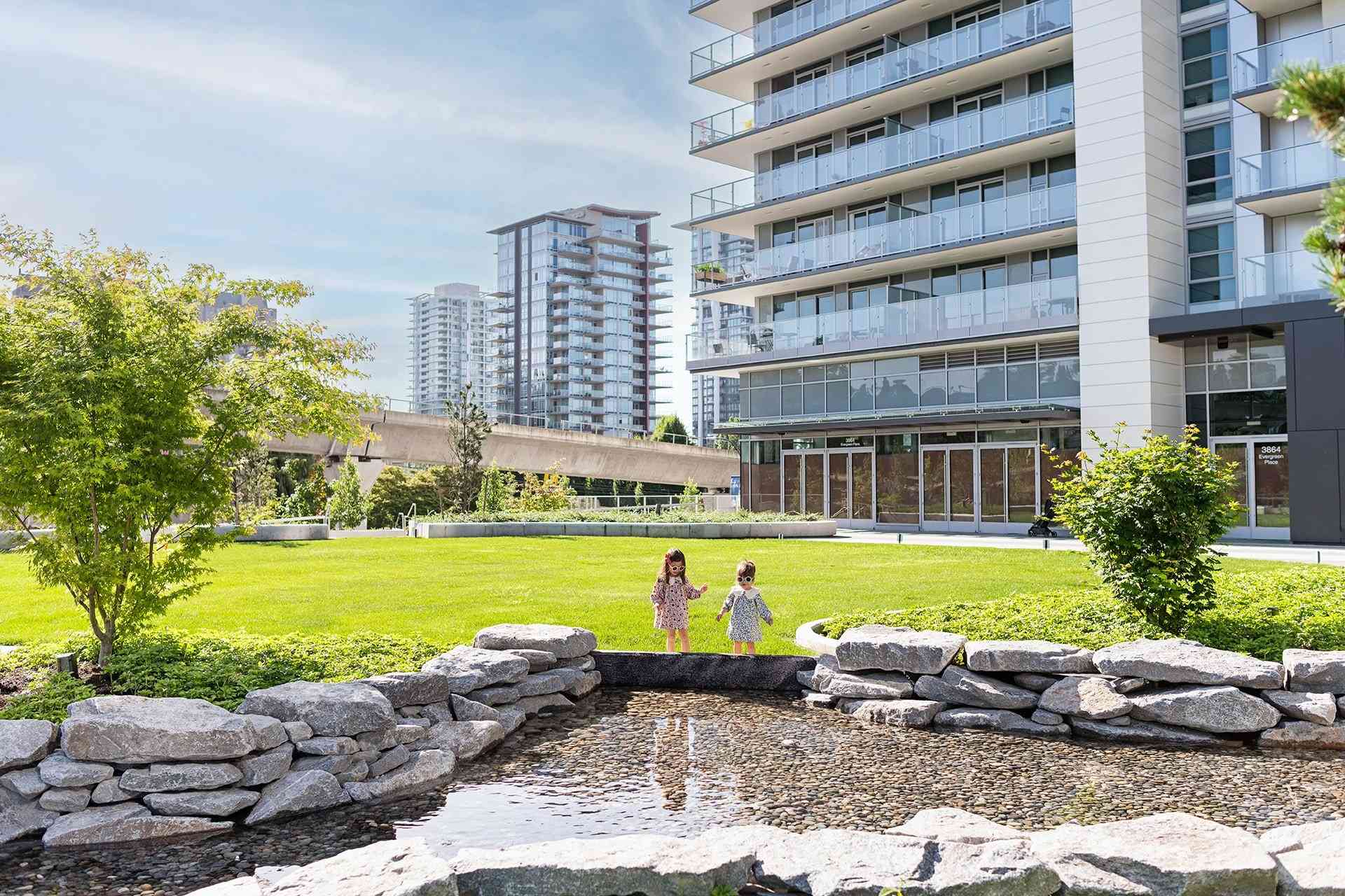The City of Lougheed – A landscaped outdoor park and courtyard area featuring a pebble-lined stone water feature, a large green lawn, and two children standing near the edge. In the background, modern residential high-rise towers and an elevated transit track rise under a bright, sunny sky.