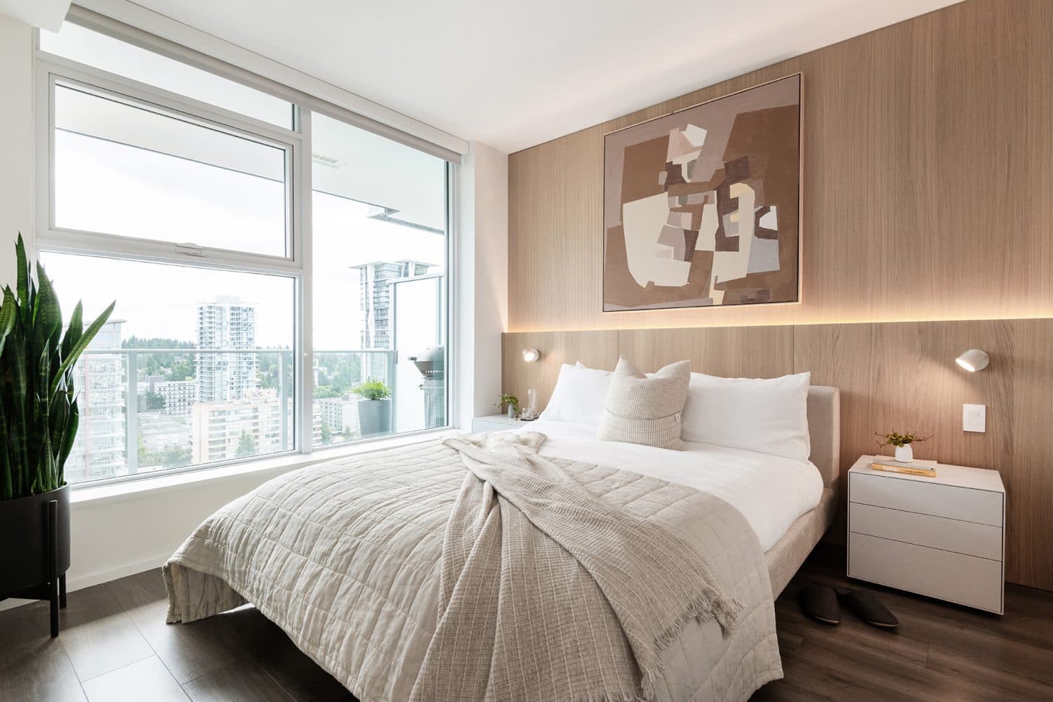 The City of Lougheed – A modern bedroom features a neatly made bed against a light wood-paneled accent wall with integrated warm backlighting, reading sconces, and abstract artwork above. Large floor-to-ceiling windows provide abundant natural light and look out toward high-rise buildings and surrounding trees.