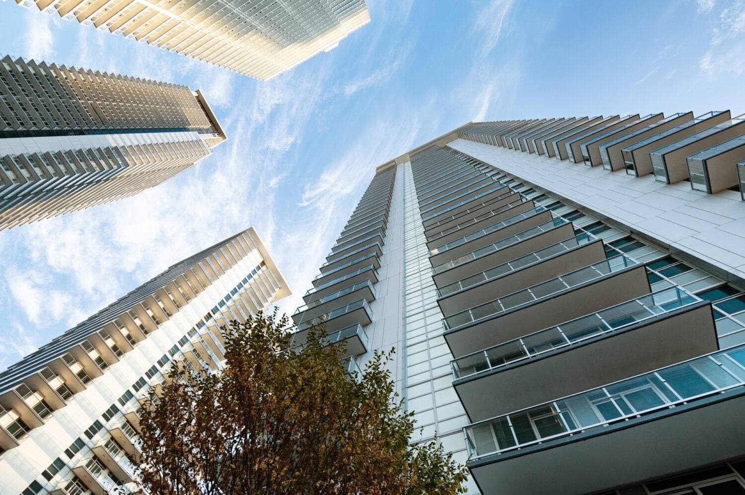 The City of Lougheed – A dynamic low-angle worm's-eye view looking straight up at multiple modern residential high-rise towers rising into a blue sky with light, wispy clouds. The buildings feature crisp white panel facades, glass balcony railings, and large windows, framed by autumn tree foliage near the ground.