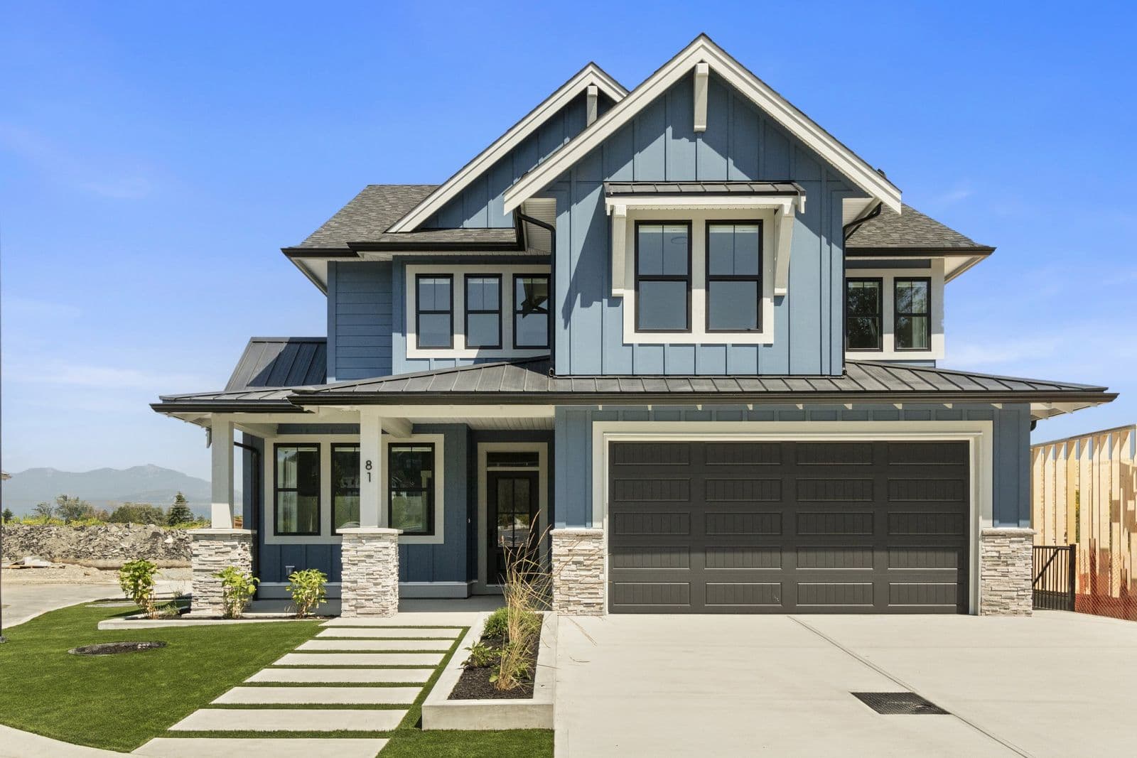 Iron Horse – Blue-gray two-story house with a covered porch, stone entry pillars, a dark double garage door, and a paved path across the front lawn, address number 81.