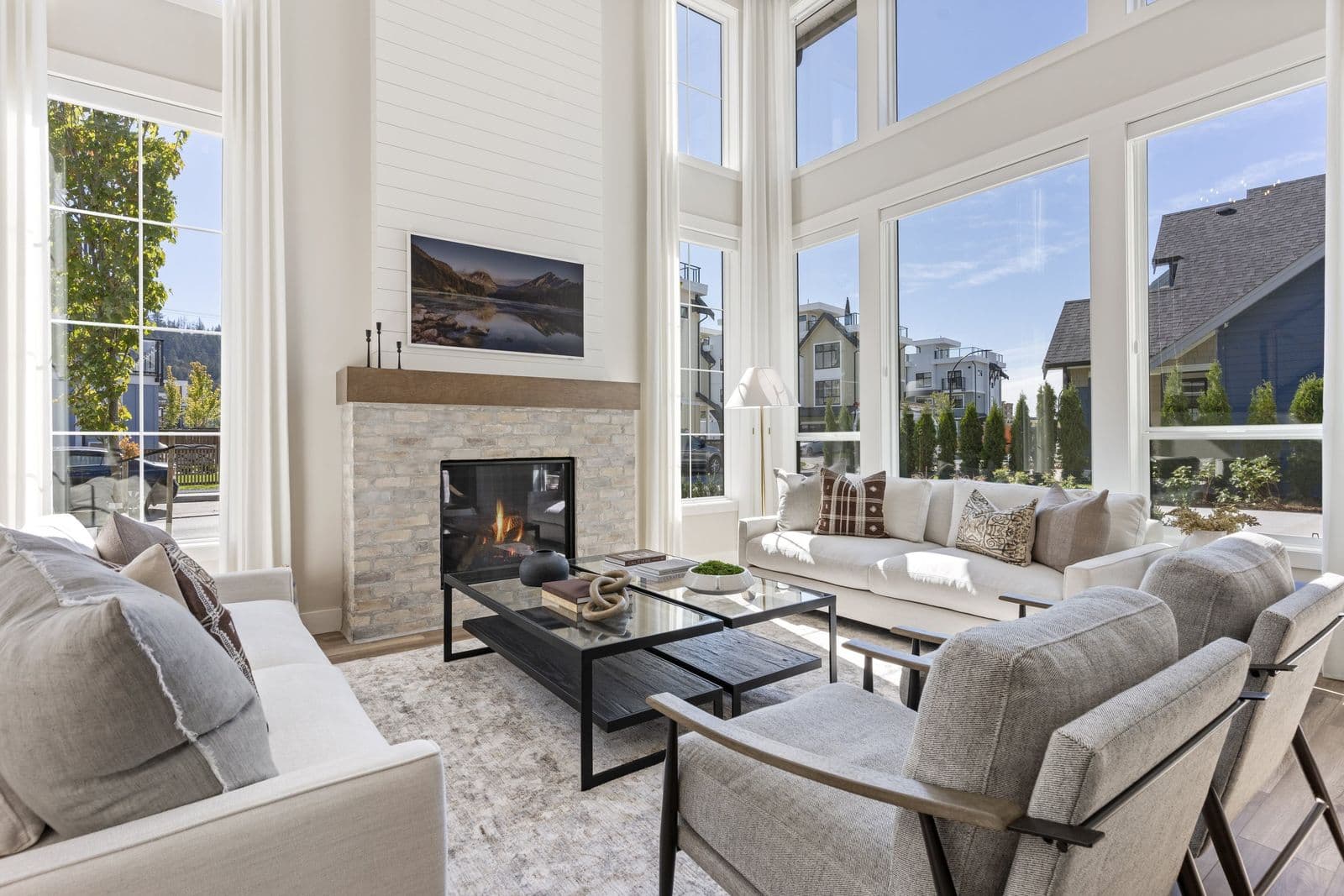 Iron Horse – Double-height living room with a stone-clad gas fireplace, mounted TV, two cream sofas and an armchair, and tall windows looking out to neighboring houses.