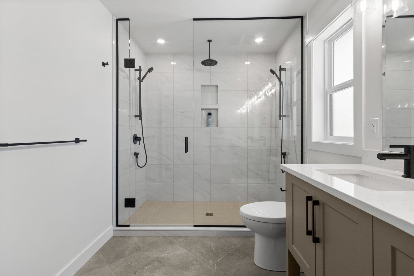 Iron Horse – Bathroom with a large glass-enclosed shower featuring marble tile and a rain shower head, a toilet, and a light wood double vanity with black fixtures.