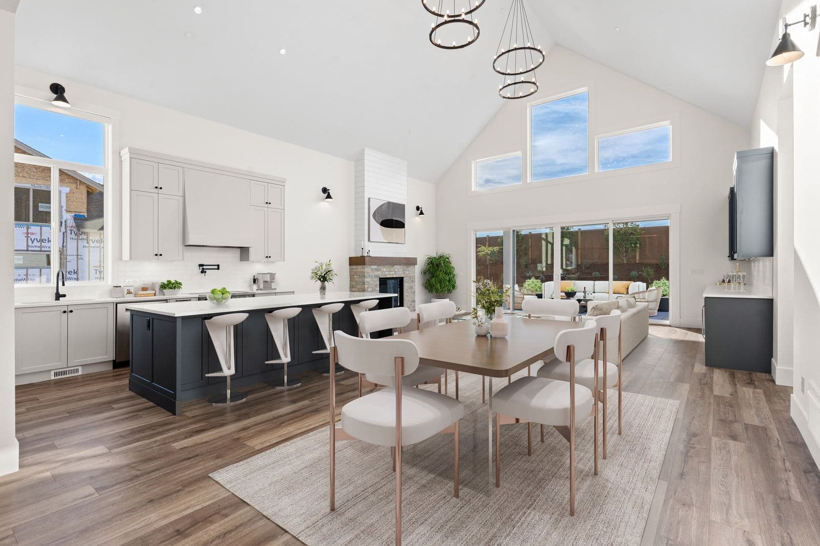 Iron Horse – Open-concept kitchen and dining area with light cabinetry, a dark blue island with white stools, a vaulted ceiling with ring pendant lights, and an unfinished house visible through the windows.