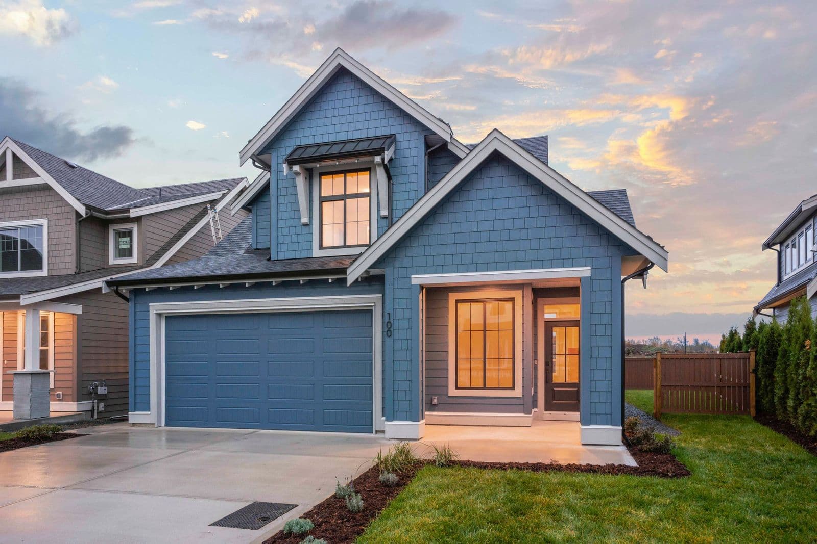 Iron Horse – Blue two-story house with an attached garage and lit windows photographed at dusk, address number 100 above the front door.