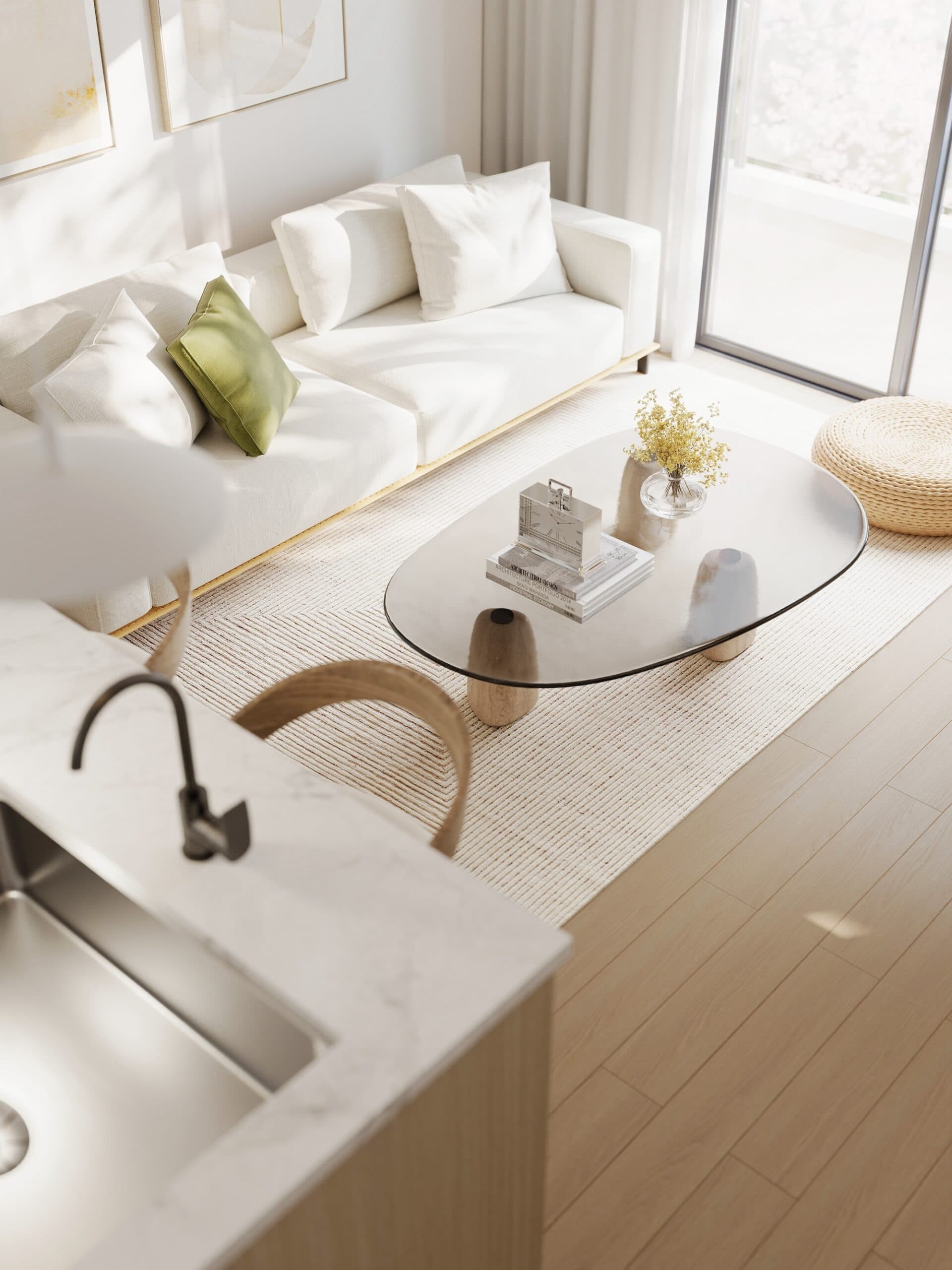 Kamilia – A high-angle view looking down into a sunlit open-concept living area, featuring a white upholstered sofa accented with a green throw pillow and an oval glass-topped coffee table over a neutral area rug. In the foreground, a kitchen island displays a marble countertop and a modern dark faucet, while large glass sliding doors let in ample daylight.