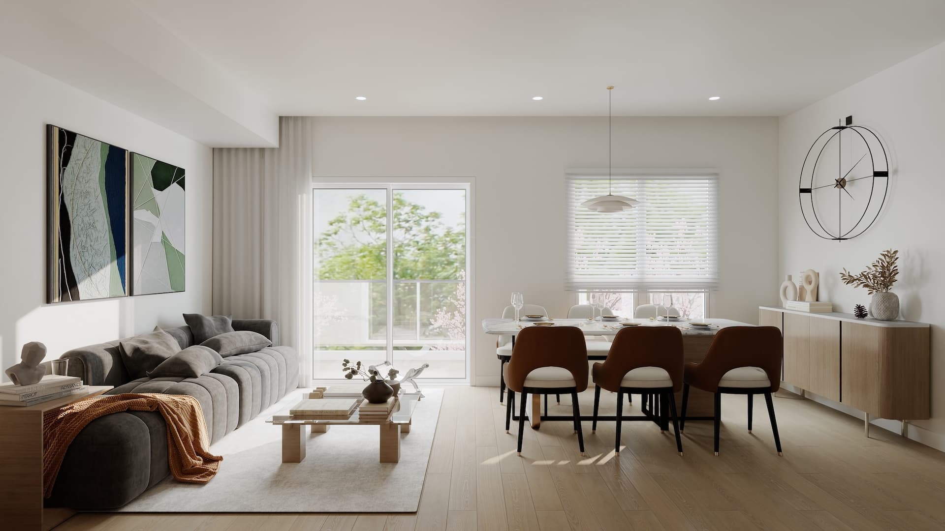 Kamilia – A bright, open-concept living and dining room featuring light hardwood floors, a plush grey channel-tufted sofa, and a dining table set with modern brown and white chairs. Sliding glass doors and a window with white blinds allow abundant daytime light to enter, looking out toward lush green trees.
