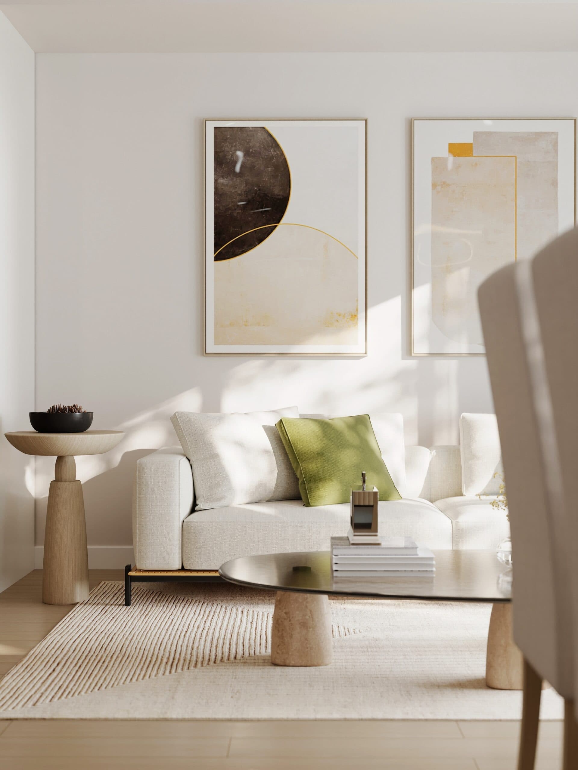 Kamilia – A sunlit, minimalist living room featuring a low-profile white upholstered sofa accented with a green throw pillow beneath two large framed abstract art prints. The space is styled with a light textured area rug, a sculpted wooden pedestal side table, and an oval coffee table with stone column legs under soft, dappled natural light.