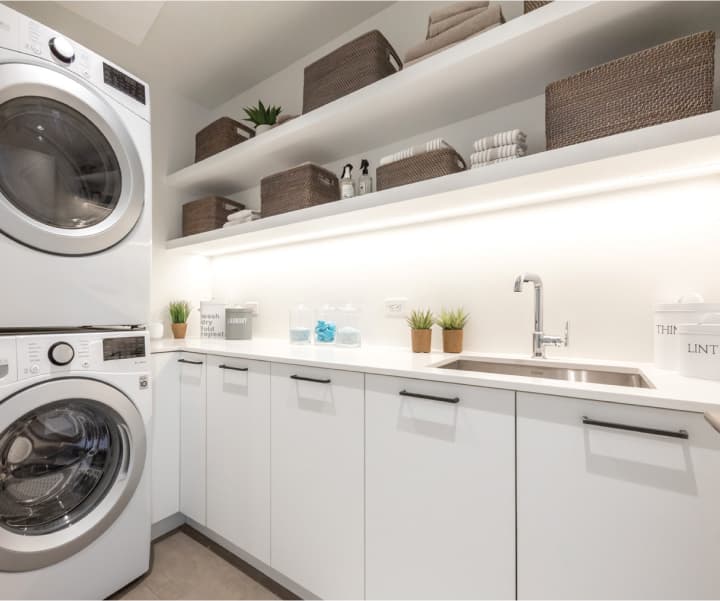 Foster Martin – A modern laundry room equipped with a stacked front-loading washer and dryer alongside an L-shaped white countertop with an undermount stainless steel sink. Open floating shelves above feature integrated under-shelf lighting and are neatly styled with woven storage baskets, folded towels, and small potted plants.