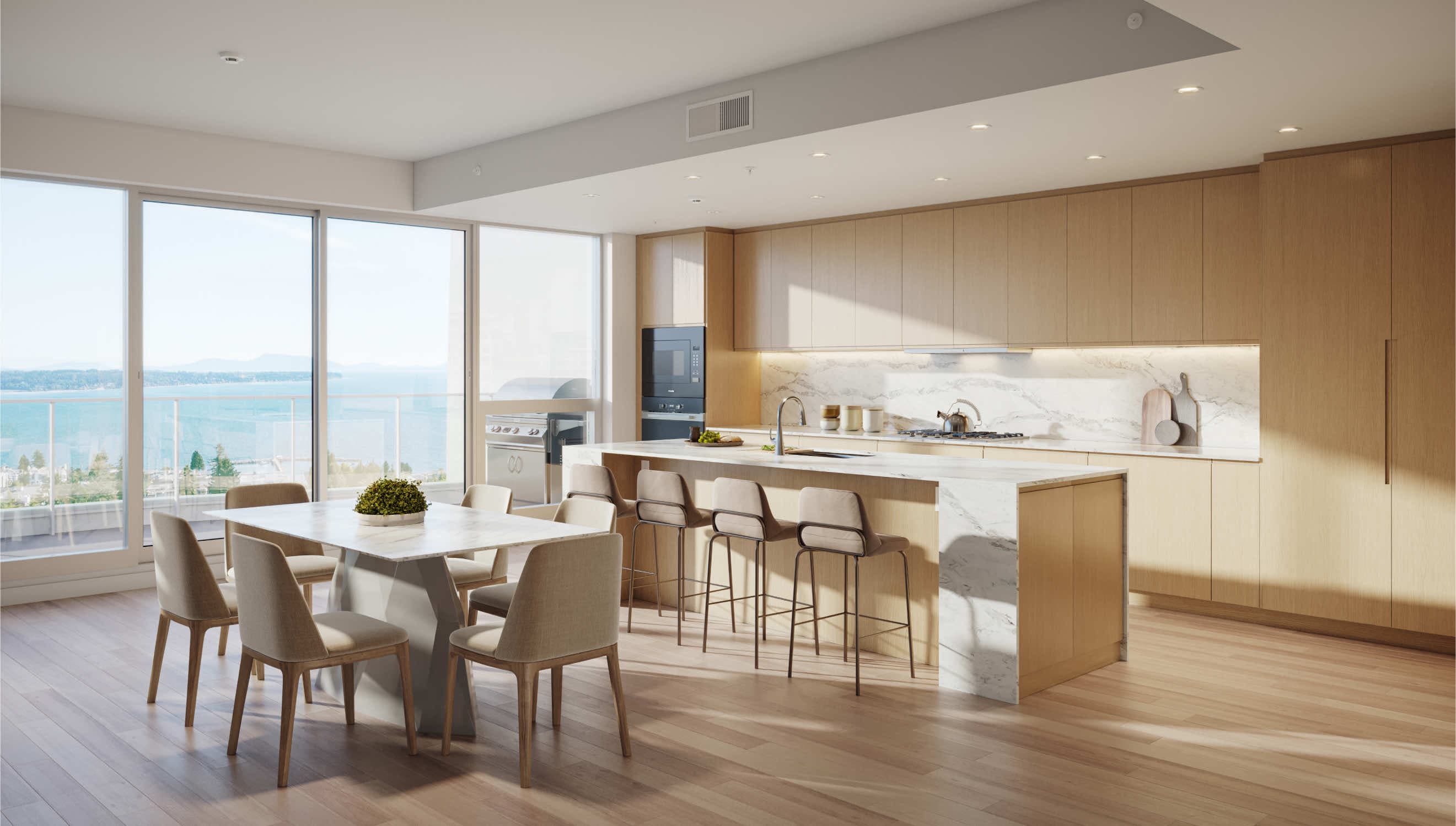 Foster Martin – A bright, open-concept kitchen and dining area features light wood flat-panel cabinetry, a waterfall marble island with barstools, and an adjacent marble dining table. Floor-to-ceiling glass doors open to an outdoor balcony with a barbecue grill and expansive views of the ocean under a clear sky.