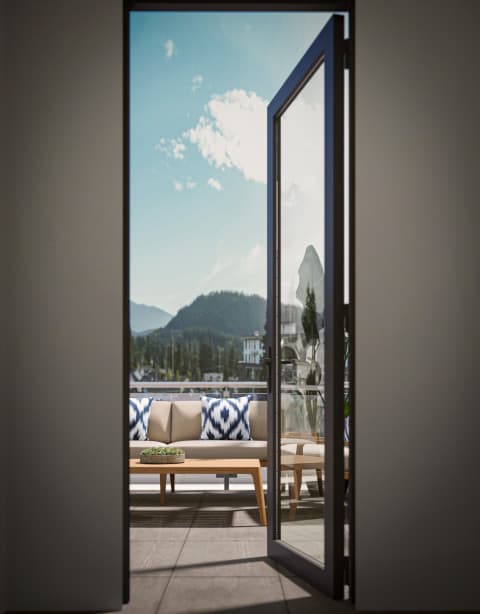 Burqville – A rooftop terrace seen through an open glass door, with an outdoor sofa and patterned cushions, a coffee table, and a mountain view beyond the railing.