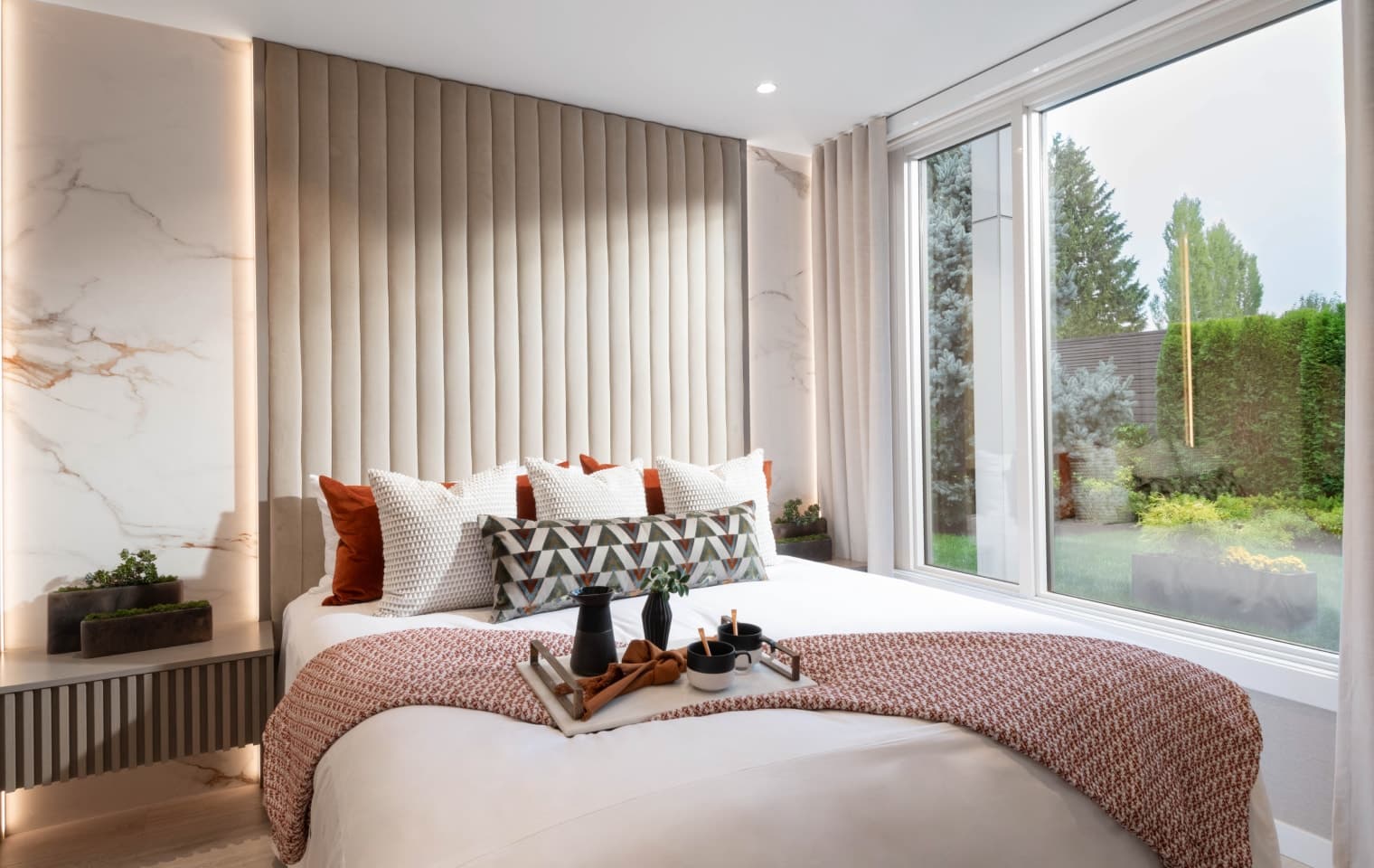 Talistar – A modern bedroom features a styled bed with a textured throw blanket, patterned accent pillows, and a breakfast tray set near a floor-to-ceiling glass window. The headboard wall includes floor-to-ceiling vertical channel padding flanked by marble-look panels with warm integrated backlighting, overlooking a lush green garden outside.