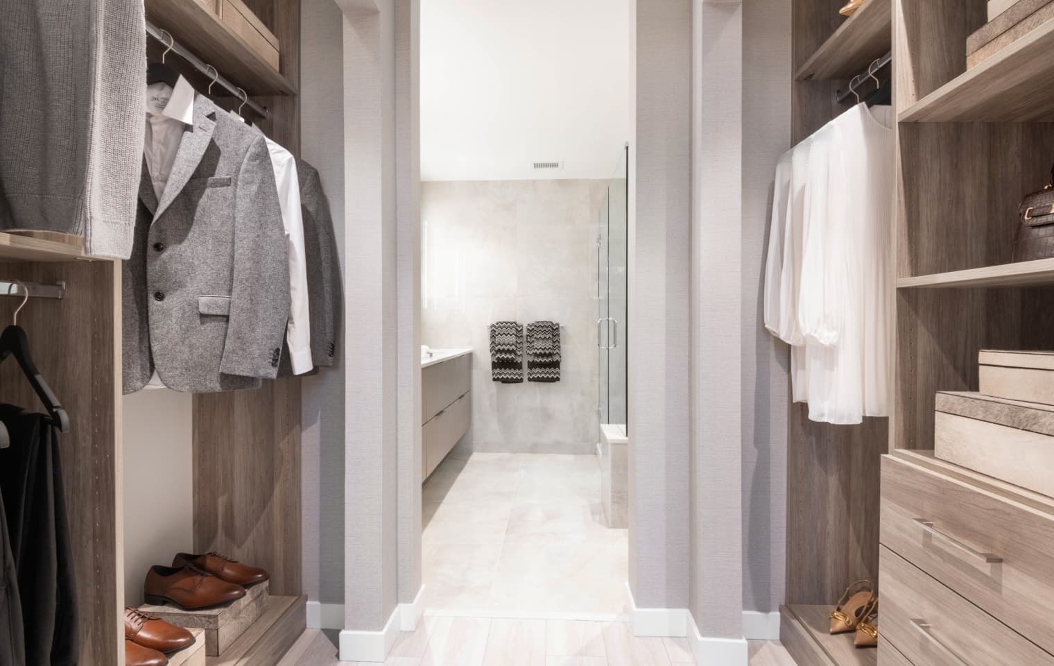Talistar – A view looking through a custom walk-in closet into an ensuite bathroom, featuring organized light-wood built-in shelving with clothing, shoes, and storage boxes. The bright bathroom beyond showcases large beige wall tiles, a floating vanity, and a glass-enclosed walk-in shower.