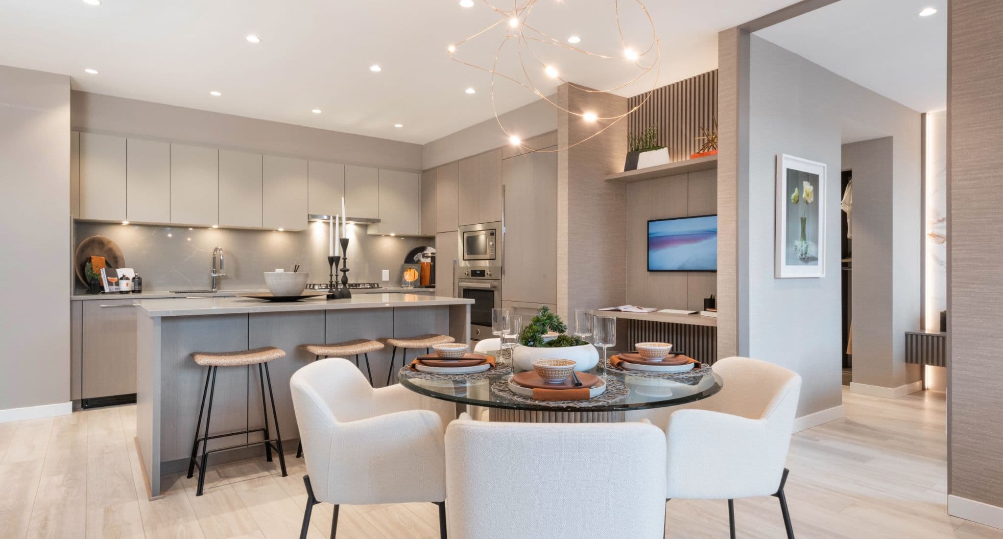Talistar – A modern open-concept kitchen and dining area features sleek, taupe flat-panel cabinetry, integrated appliances, and a large kitchen island with counter seating. In the foreground, a round glass dining table set for four with white upholstered chairs is illuminated by a delicate wire orb chandelier.