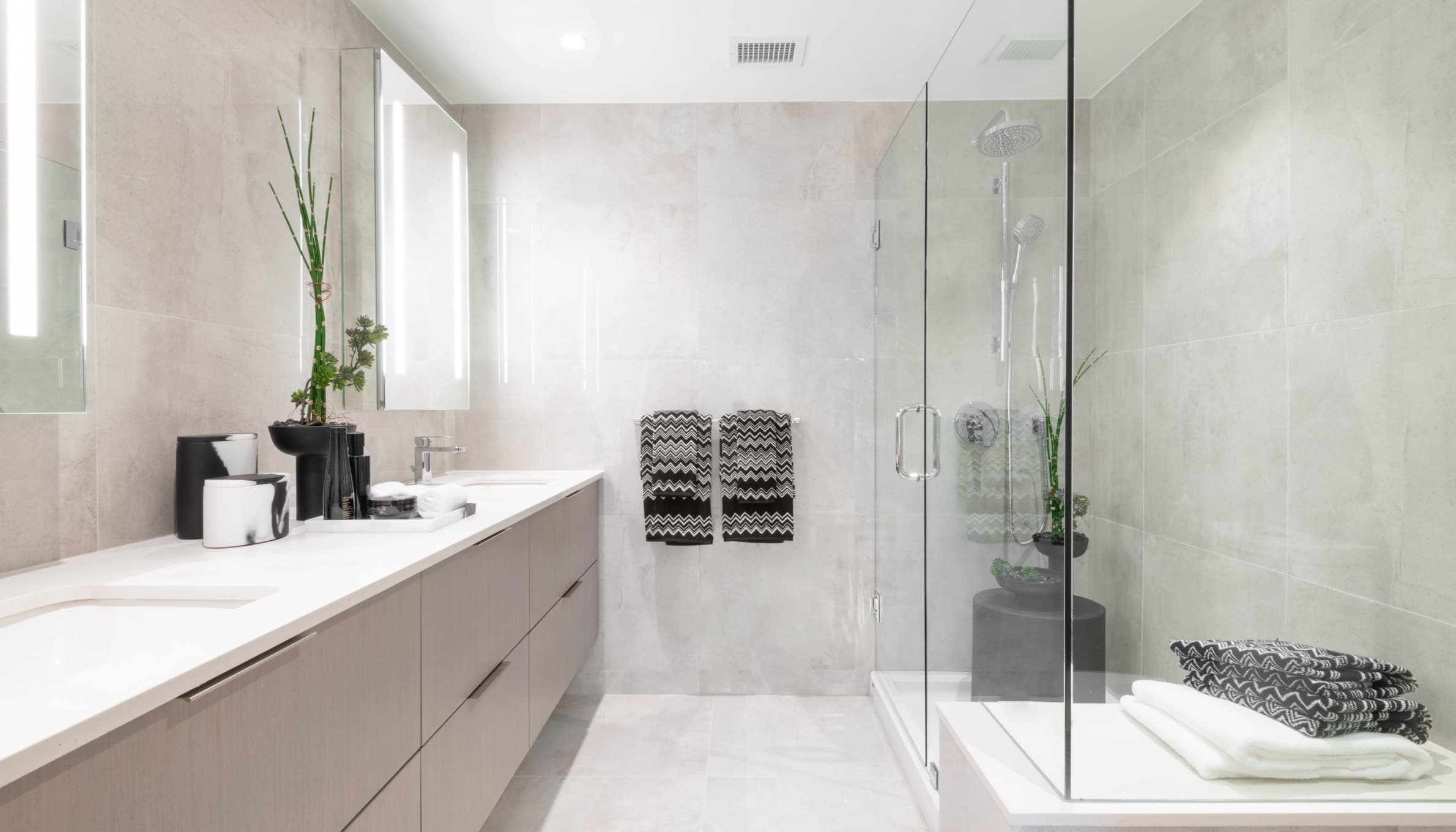 Talistar – A modern, spa-inspired bathroom featuring a long light-wood floating vanity with white countertops, integrated sinks, and back-lit mirrors. Light gray large-format tiles cover the floor and walls, complementing a glass-enclosed walk-in shower equipped with polished chrome fixtures and subtle decorative accents.