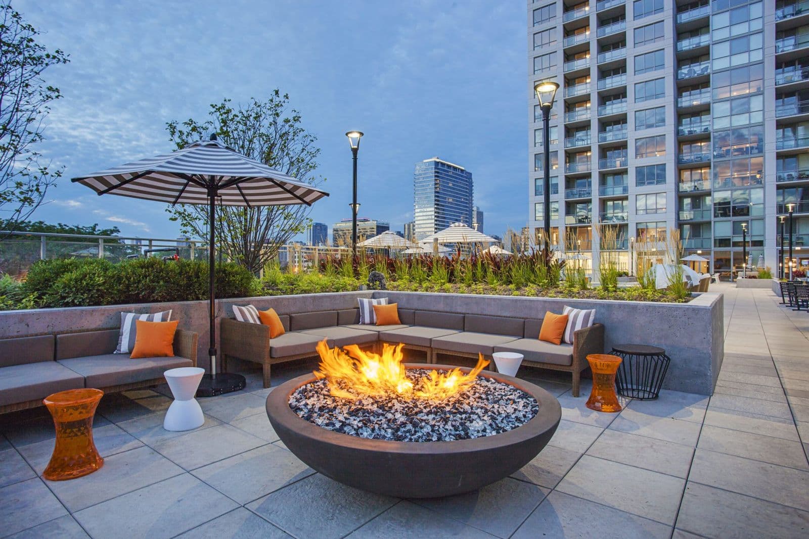 Impressions Condos – Rooftop lounge with a built-in fire pit, cushioned sectional seating with orange pillows, umbrellas, and a city skyline view at dusk.