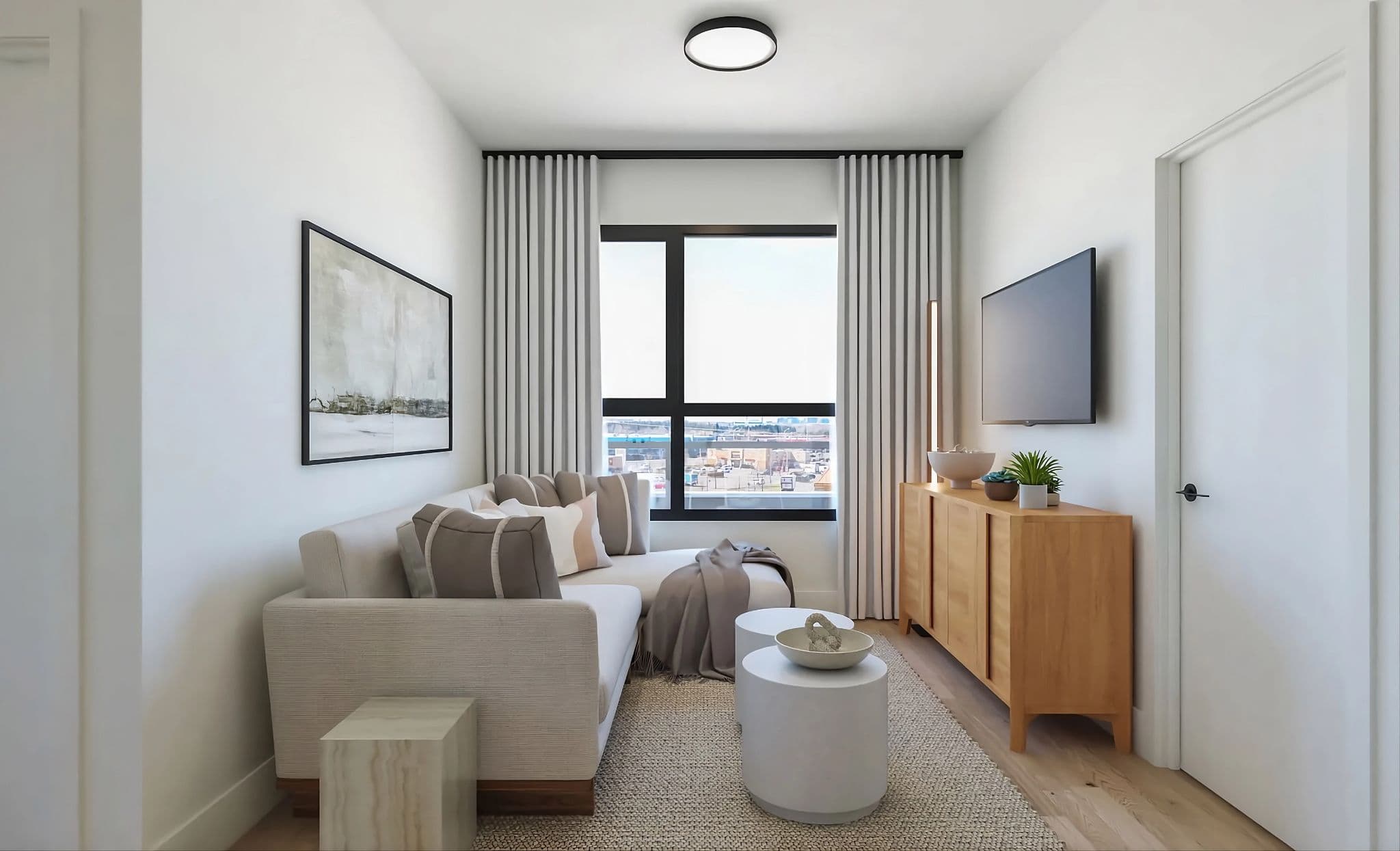 Aavand – A bright, compact living room features an L-shaped beige sectional sofa styled with neutral accent pillows and a throw blanket, placed opposite a wall-mounted flat-screen TV and wooden console. Large floor-to-ceiling drapery frames a black-trimmed window that looks out onto an urban cityscape in daytime light.
