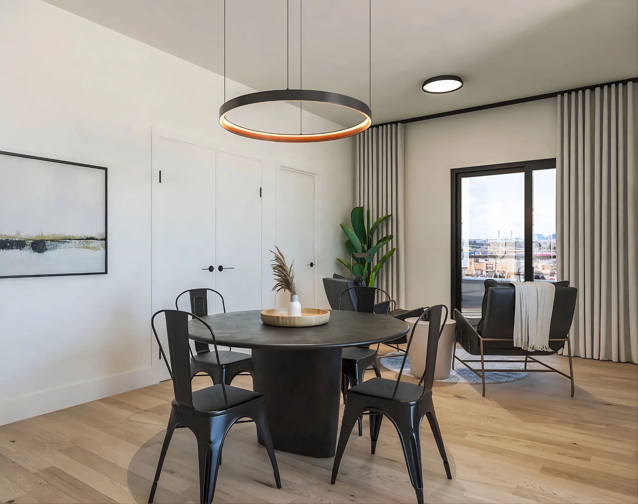 Aavand – A modern open-concept dining and living area featuring light wood flooring, a circular black dining table with matching metal chairs beneath a large halo pendant light, and a small sitting area in the background with armchairs and a potted plant. Sliding glass doors lead out to a balcony overlooking an urban cityscape under bright daylight.