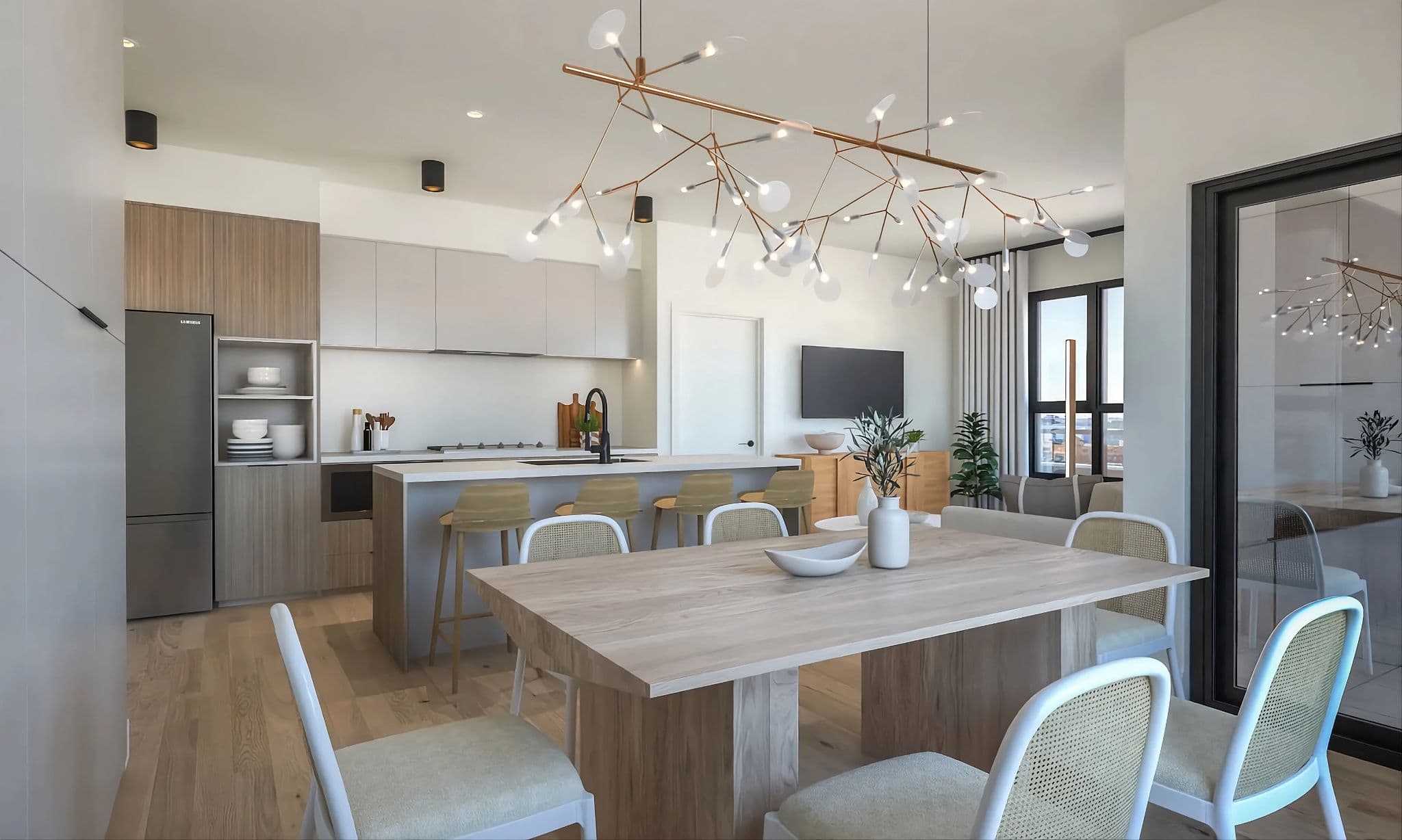 Aavand – An open-concept modern apartment interior featuring a light wood dining table with cane-back chairs beneath a striking, branch-like sculptural chandelier. In the background, the space transitions seamlessly to a kitchen island with barstools, two-tone cabinetry, and an adjoining living area with a wall-mounted TV and sliding glass doors.
