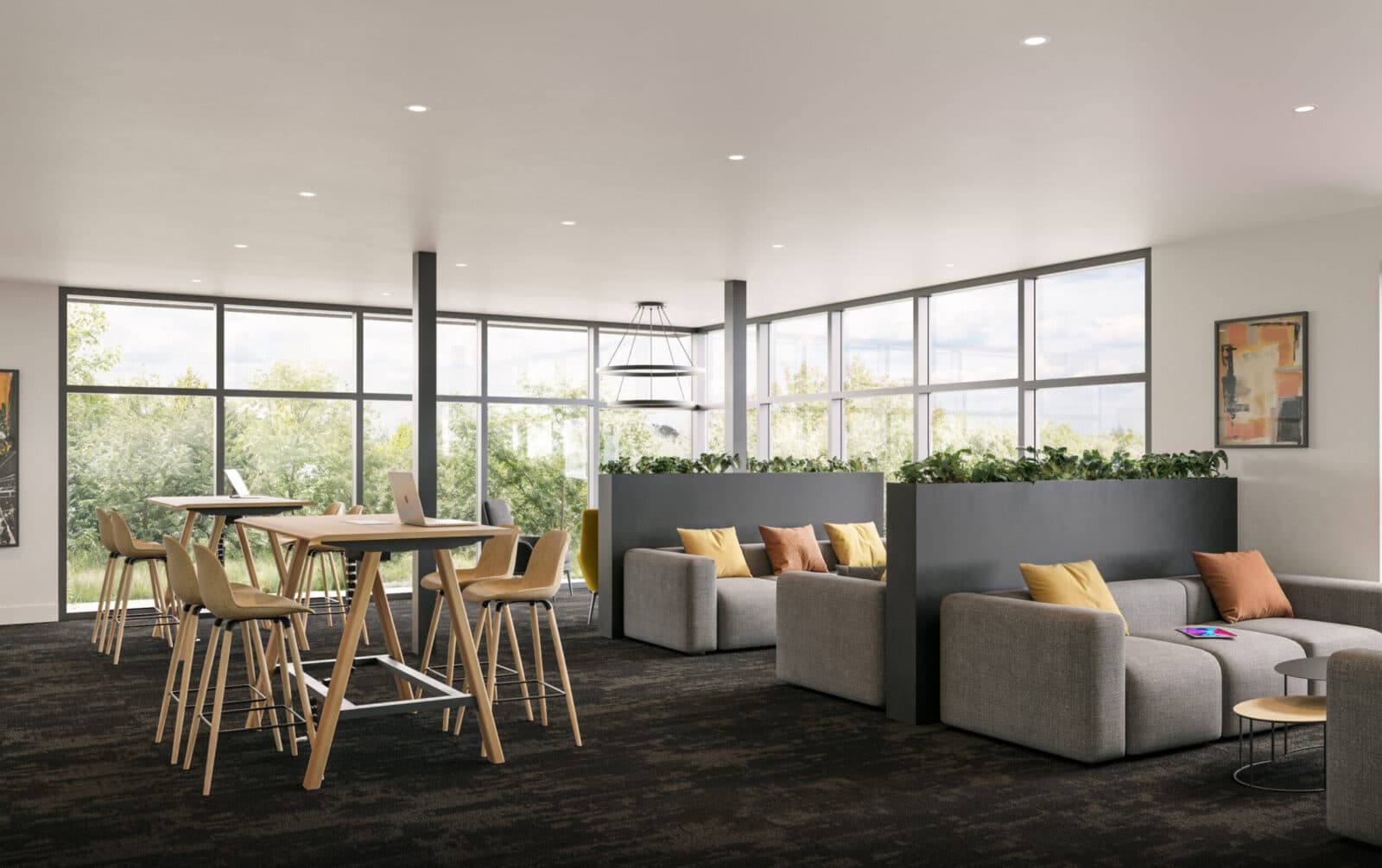 Scott + 77 – A modern co-working and lounge area with dark carpeting, high-top wooden work tables, and comfortable gray sectional sofas with yellow and orange accent pillows. Floor-to-ceiling windows line the back wall, filling the bright indoor space with natural light and views of green trees outside.