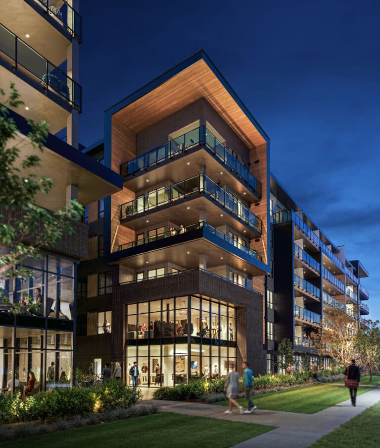 Scott + 77 – A twilight dusk view of a modern multi-story residential building featuring wood-paneled soffits, dark brick cladding, and glass balcony railings. Pedestrians walk along a paved pathway and manicured lawn beside glowing double-height ground-floor amenity spaces with floor-to-ceiling windows.