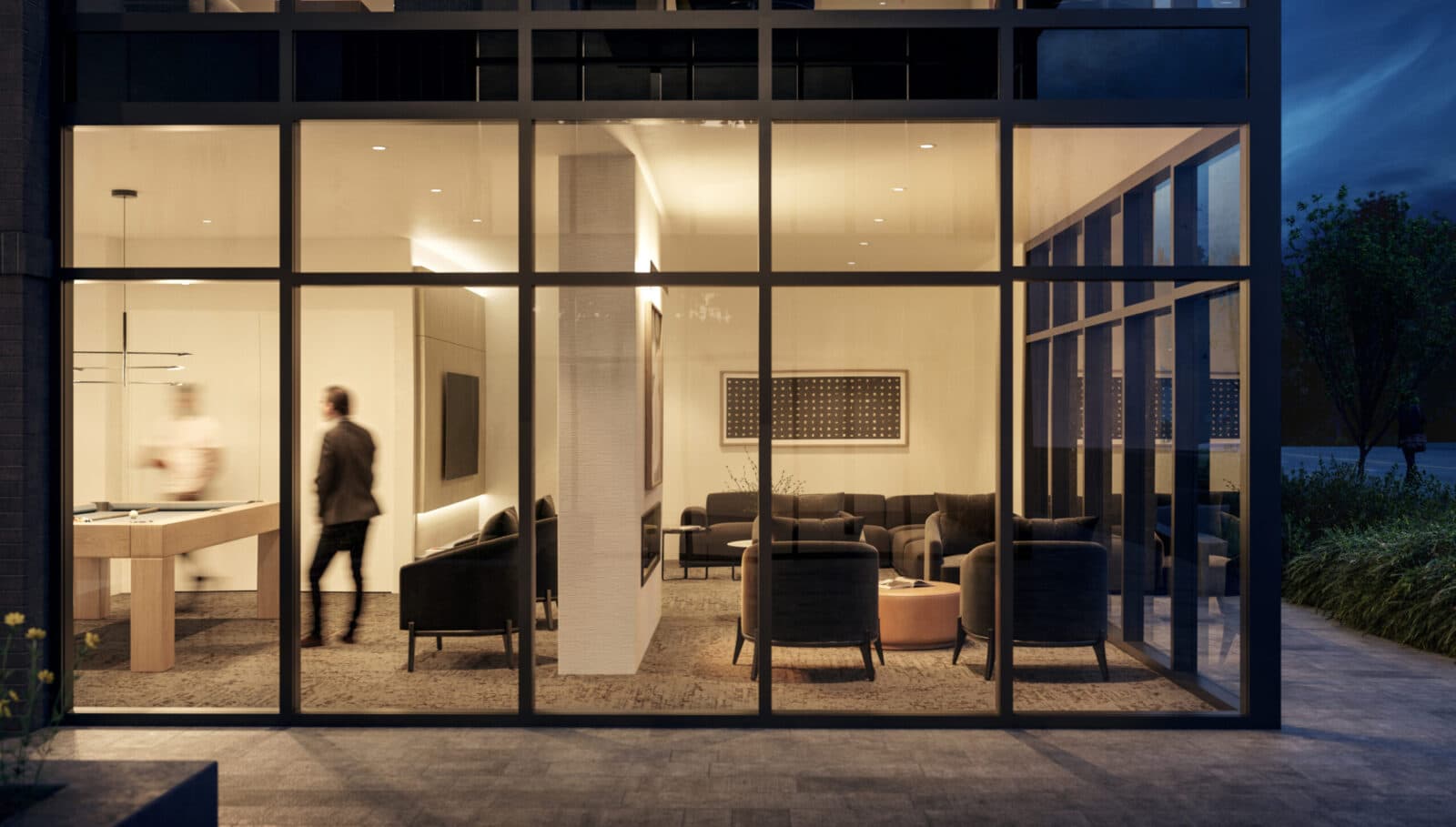 Scott + 77 – An evening street-level view looking through floor-to-ceiling glass windows into a warm, illuminated resident lounge featuring a pool table area and a plush seating section. Dark armchairs, a sectional sofa, modern artwork, and ambient recessed lighting create a sophisticated atmosphere against the dusk exterior.