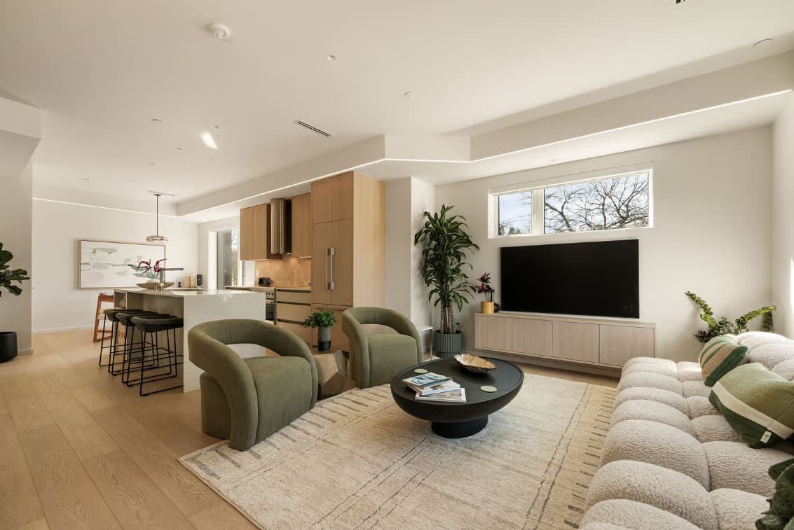 The Modern on Heather – An open-concept living and kitchen area styled with earthy green armchairs, a large textured sectional, light wood flooring, and flat-panel cabinetry. Bright natural light fills the space through a window above the wall-mounted television.
