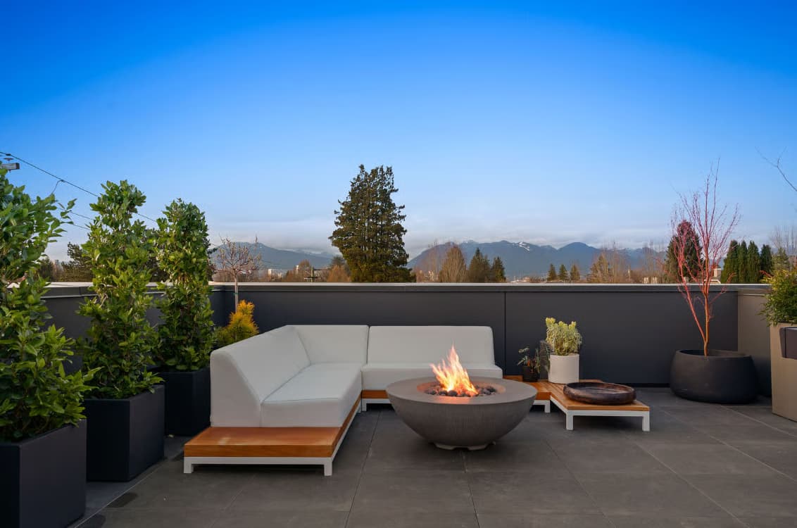 The Modern on Heather – A modern rooftop patio lounge featuring a white sectional sofa, wood accent bases, potted greenery, and a burning concrete fire bowl. The open-air space overlooks a clear blue sky and a distant mountain skyline during dusk.