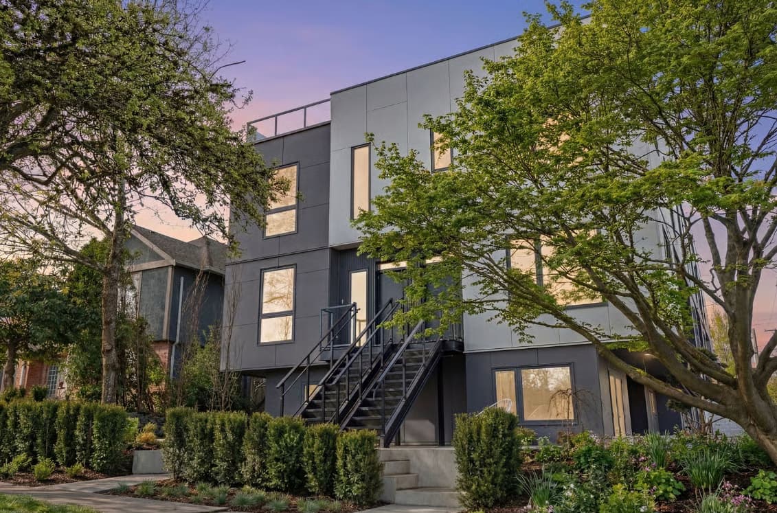 The Modern on Heather – A modern multi-story residential building featuring two-tone light grey and dark charcoal cladding, framed by lush green trees and manicured hedges at dusk. Black metal stairs lead up to the elevated main entrances, while warm interior lighting glows through the windows against a soft purple and orange sunset sky.
