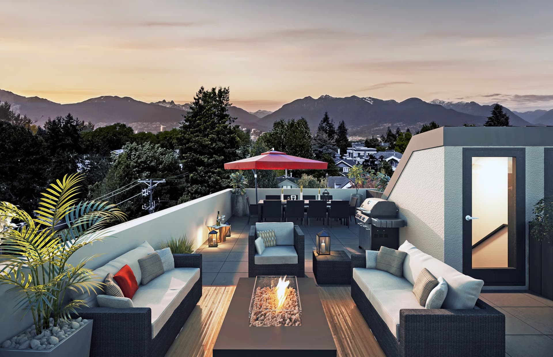 The Modern on Heather – A rooftop patio features a lounge area with dark wicker sofas centered around a lit gas fire pit, leading to an outdoor dining table shaded by a red umbrella and a stainless steel BBQ grill. In the background, lush green trees and distant, snow-capped mountains are set against a soft sunset sky.